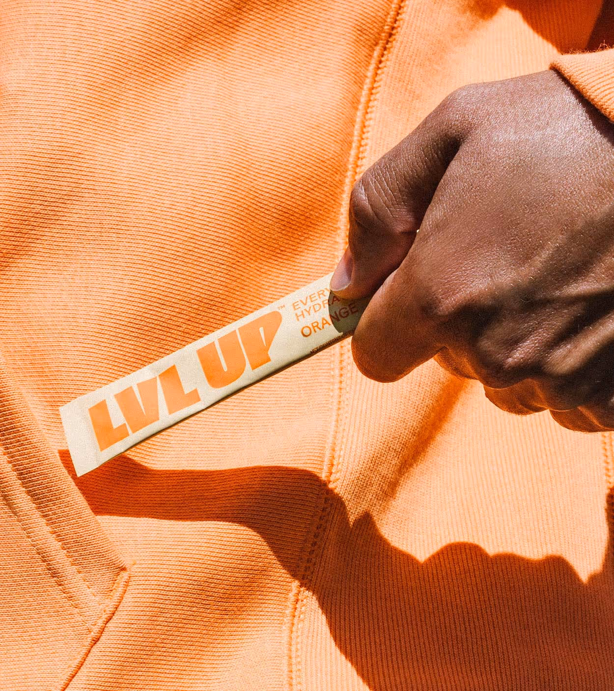 A hand holding an orange LVL UP hydration stick against an orange fabric background.