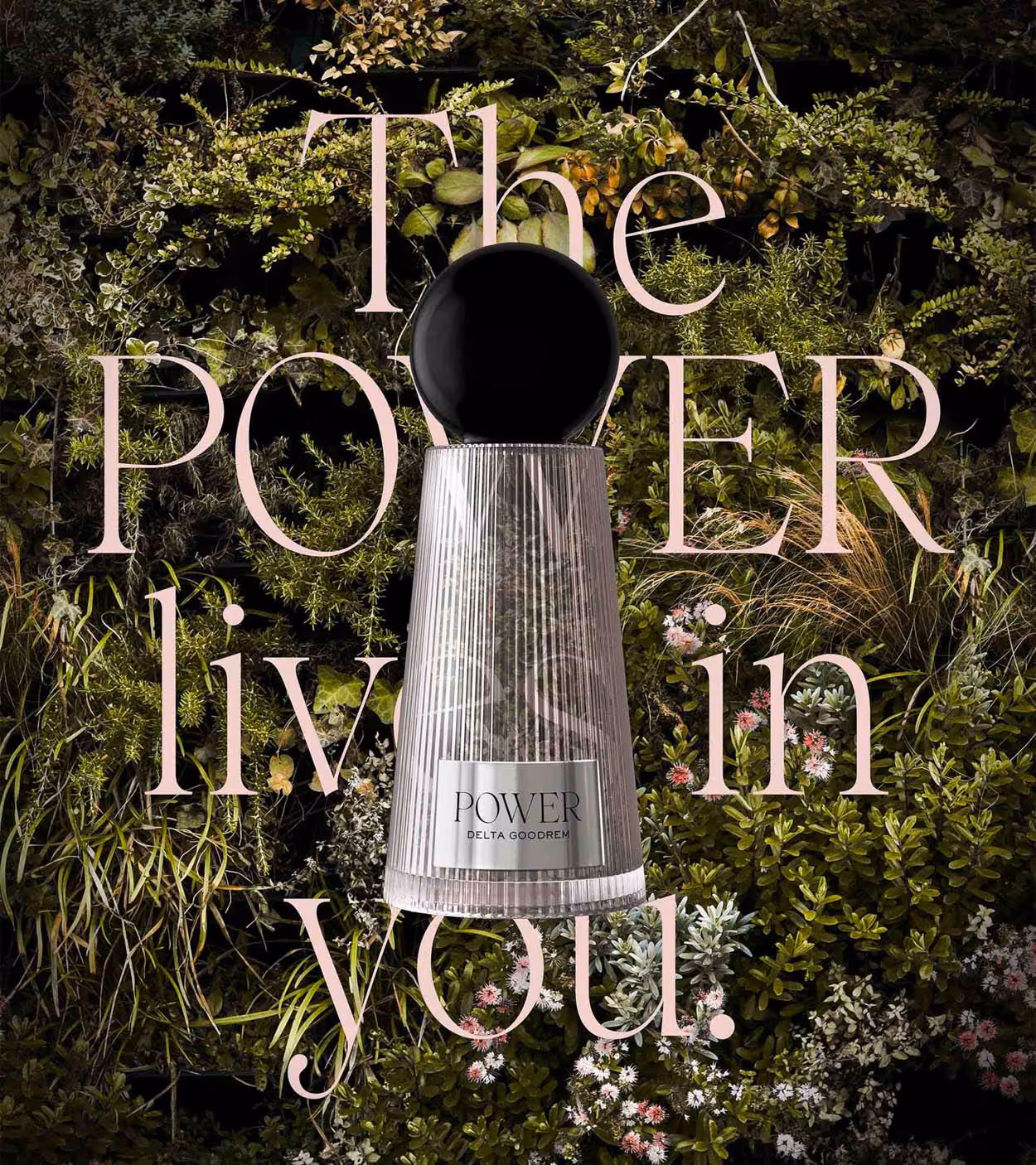 Bottle of POWER perfume by Delta Goodrem with a black spherical cap against a background of green foliage and pink flowers overlaid with text reading 'The POWER lives in you.'