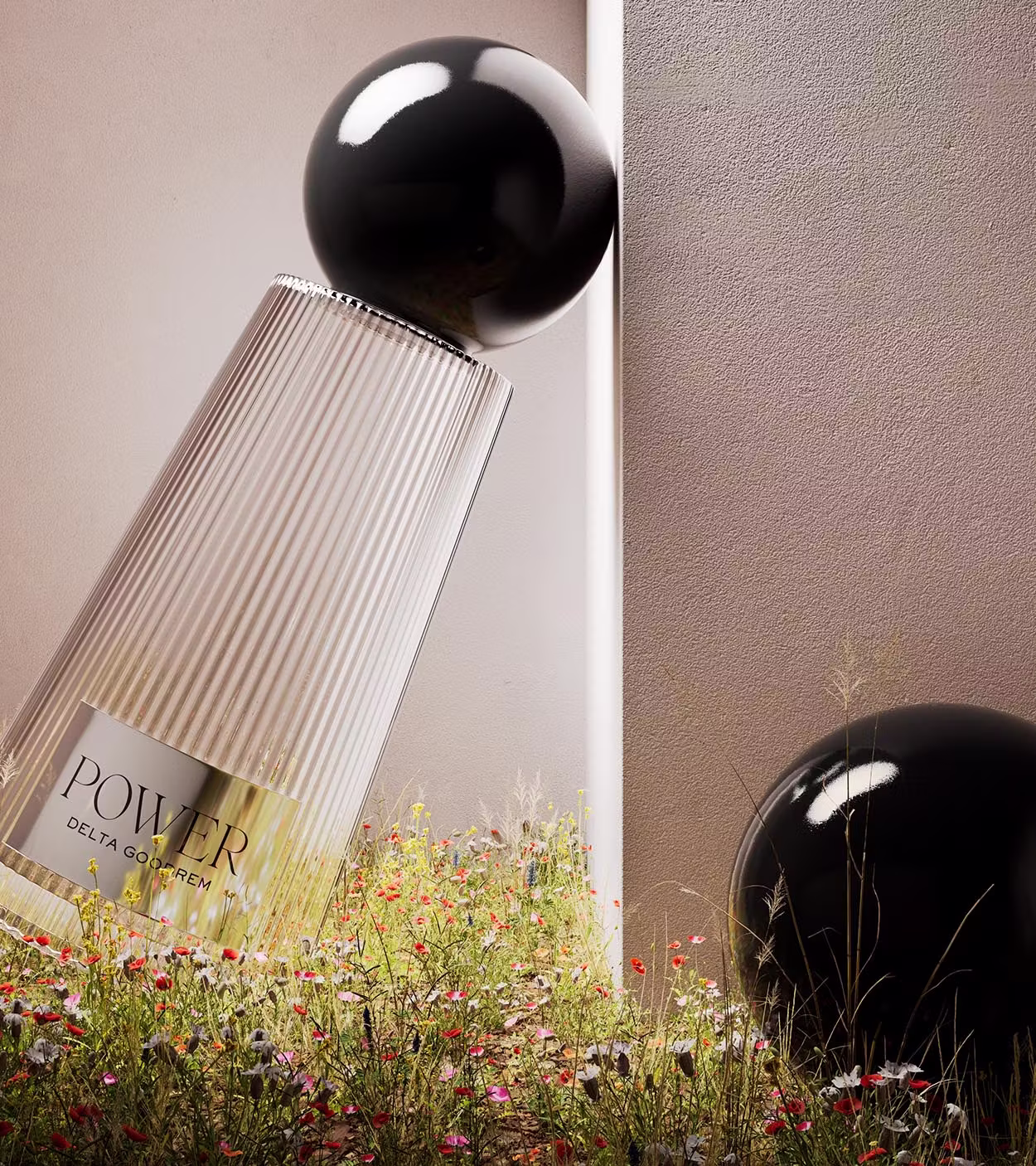 Tilted translucent perfume bottle with black spherical cap labeled 'POWER DELTA GOURREM' surrounded by wildflowers against a beige wall.