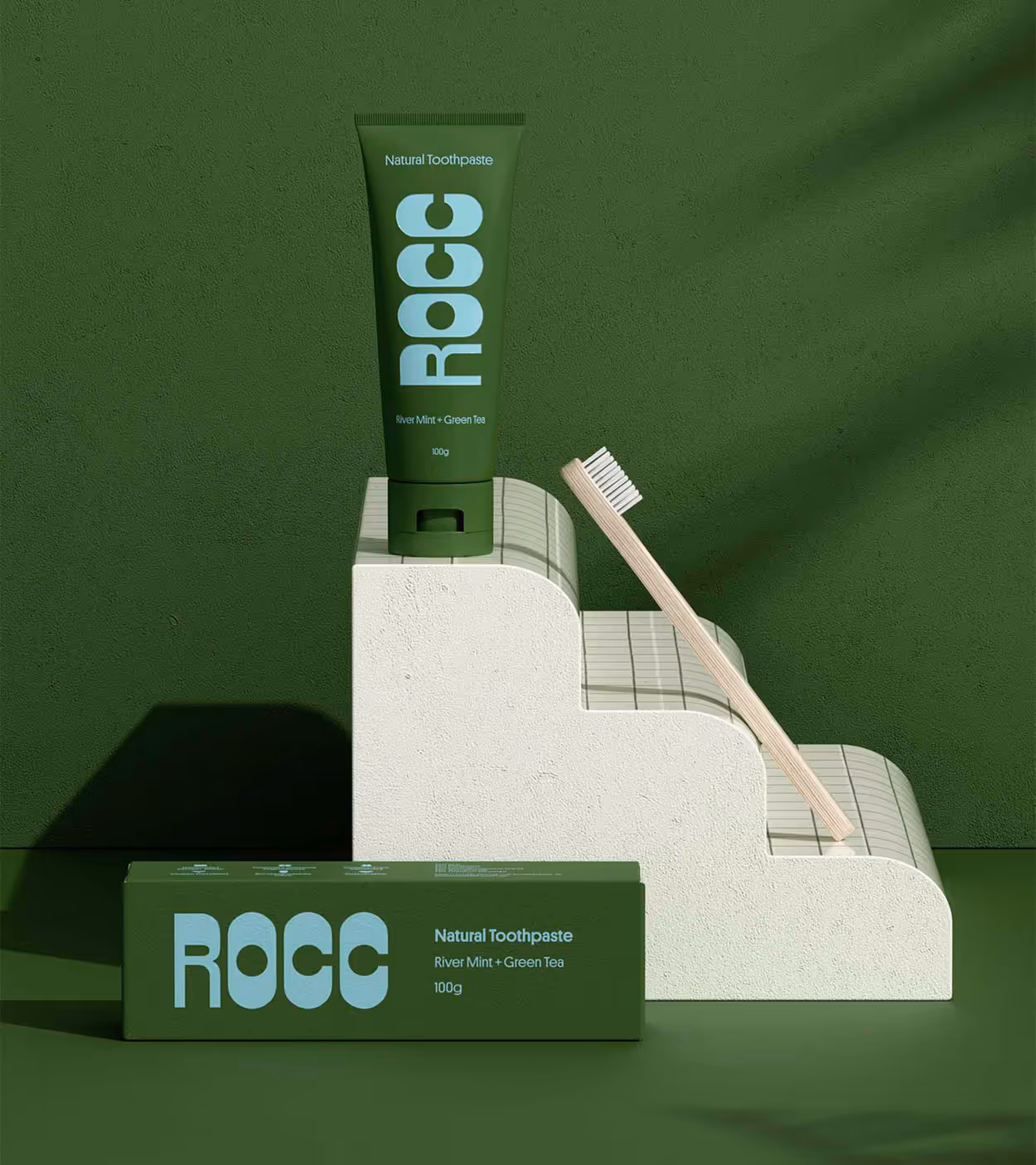 Green tube of ROCC natural toothpaste with river mint and green tea flavor, a matching box, and a wooden toothbrush on a textured white stepped platform against a green background.