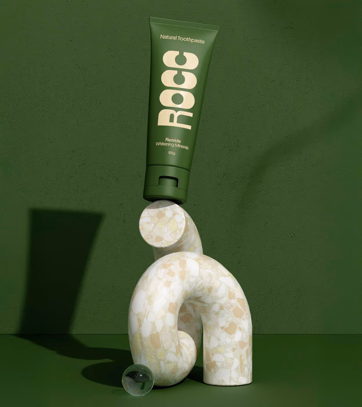 Green tube of ROCC natural toothpaste balanced on a curved terrazzo sculpture with a small transparent glass sphere on a green surface and background.