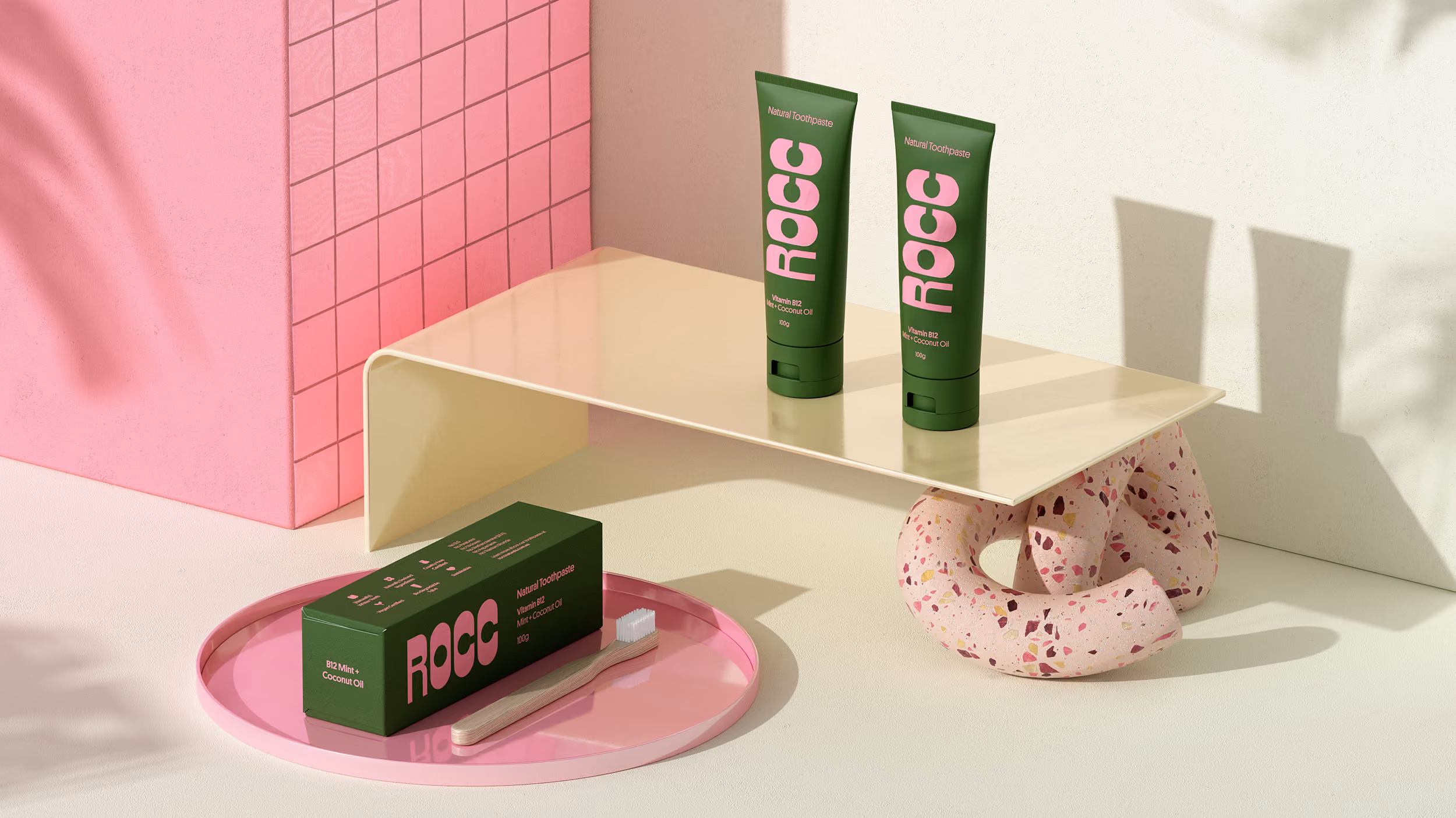 Two green tubes of ROCC natural toothpaste on a beige shelf supported by a pink terrazzo ring, with a green ROCC toothpaste box and a wooden toothbrush on a pink round tray nearby.
