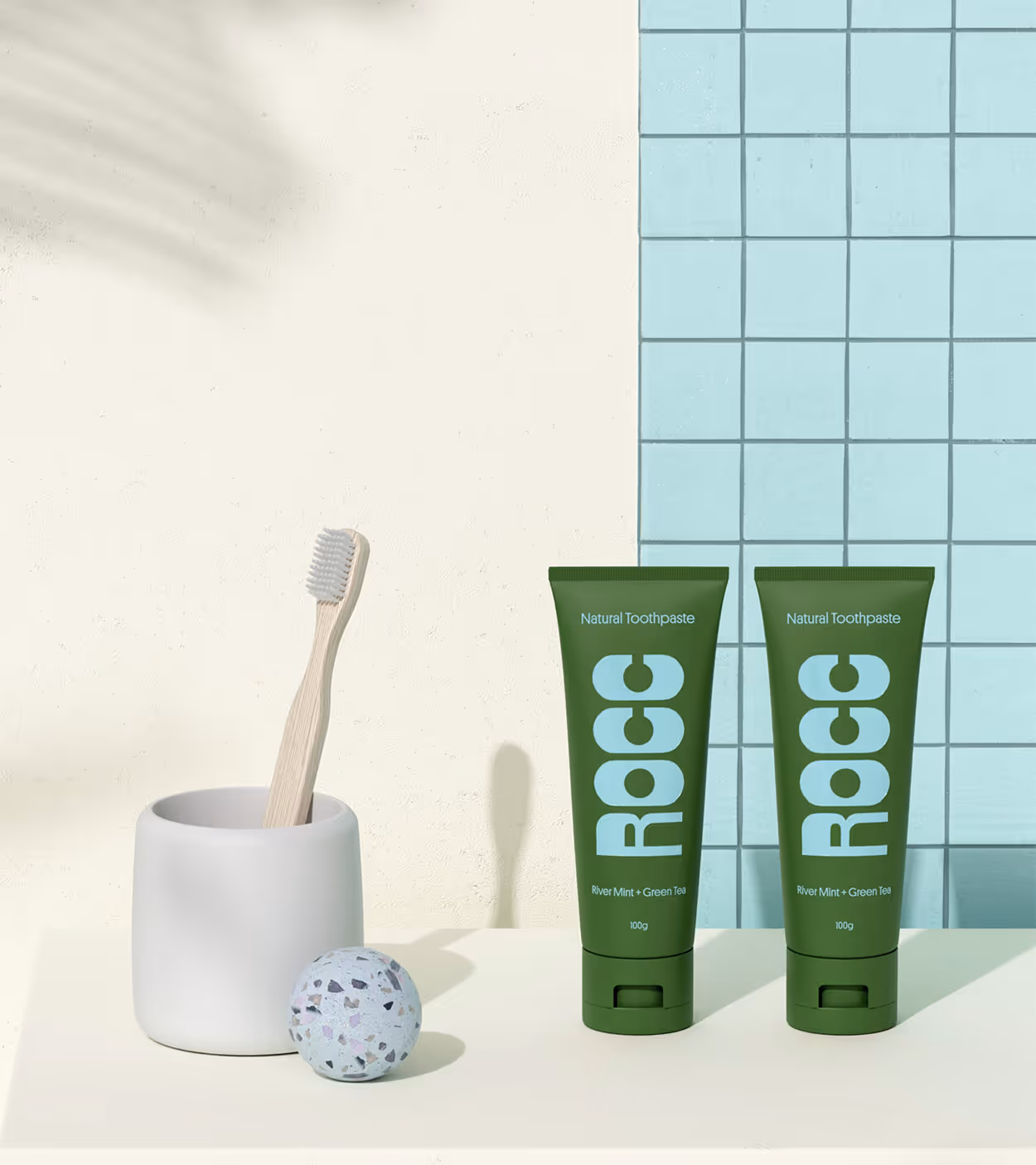 Two green tubes of ROCC natural toothpaste with river mint and green tea next to a wooden toothbrush in a white holder and a speckled bath bomb ball on a cream surface with blue tiled background.