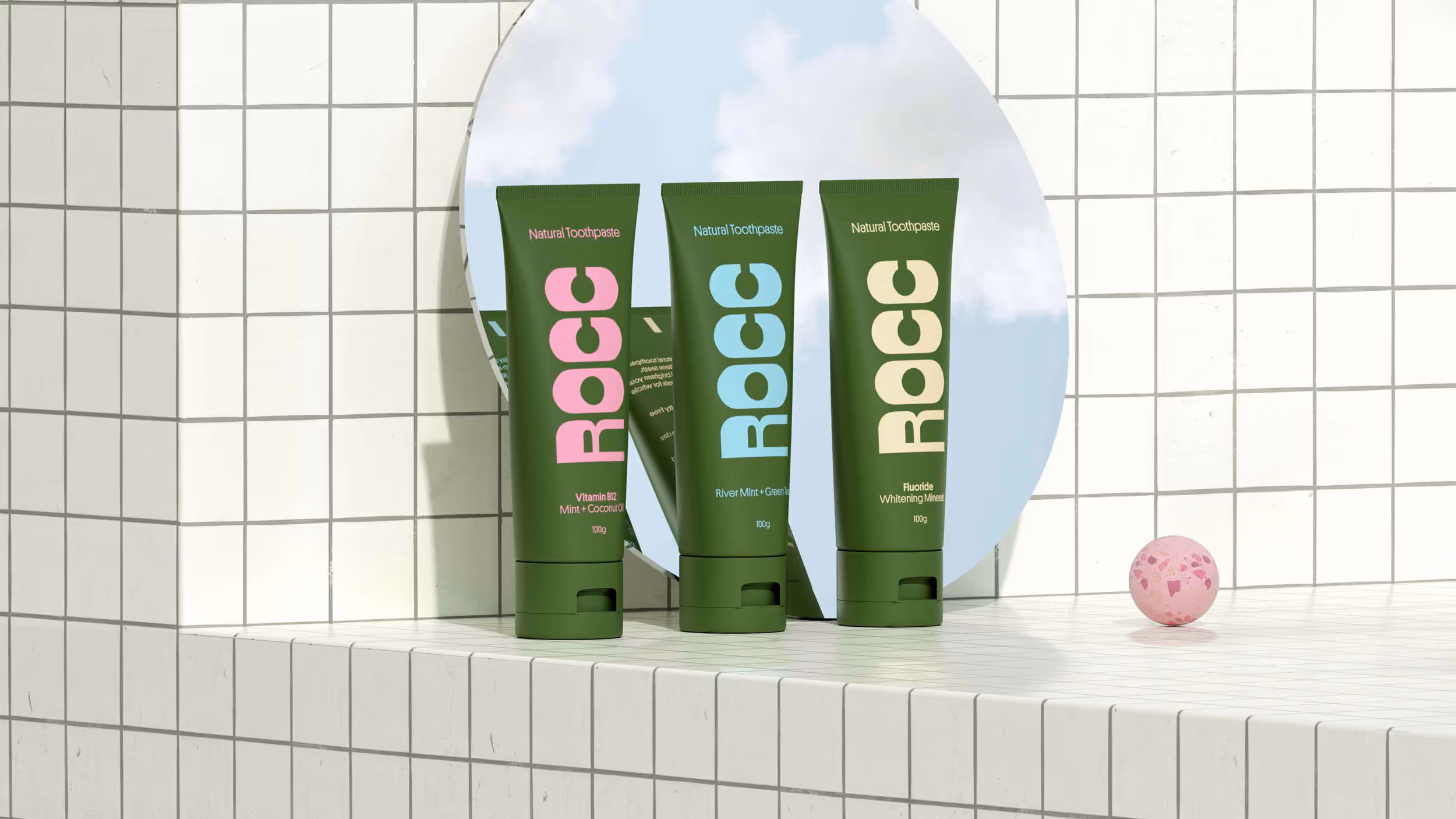 Three green tubes of ROCC natural toothpaste in pink, blue, and beige text, displayed in front of a round mirror reflecting blue sky with clouds, on tiled white surface with a pink speckled sphere.