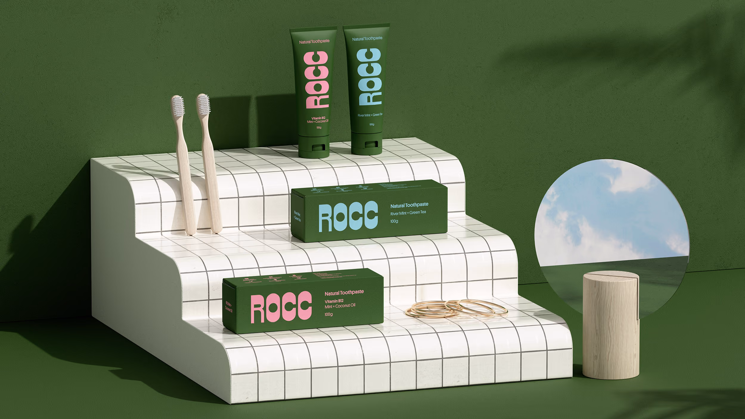Two natural toothpaste tubes and two matching toothpaste boxes labeled ROCC displayed on white tiled stepped platform with two wooden toothbrushes and a round mirror reflecting a cloudy sky on a green background.