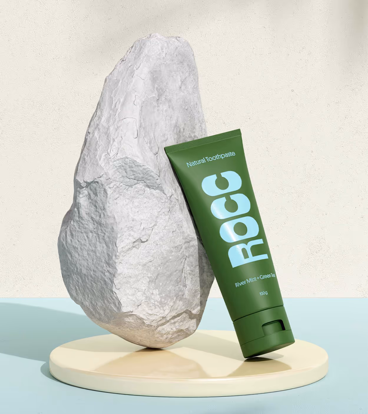 Green tube of Rocc natural toothpaste leaning against a large textured white rock on a cream circular platform with a light blue background.