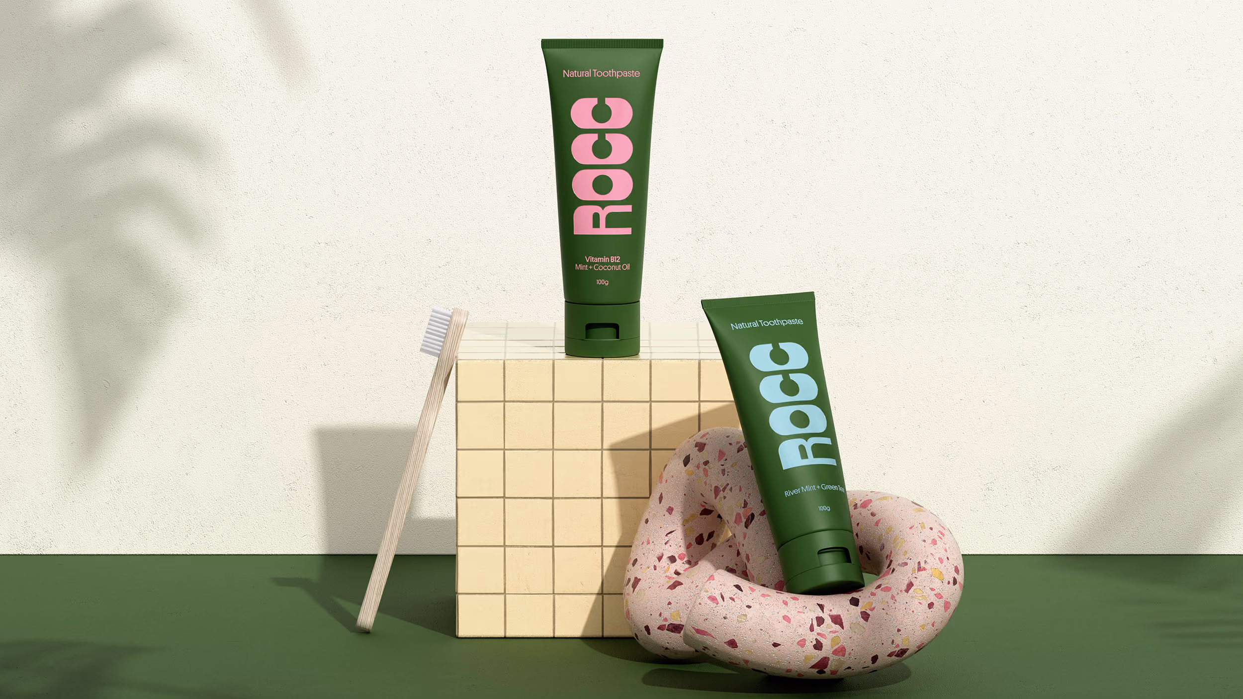 Two green tubes of ROCC natural toothpaste, one standing on a tiled beige cube with a wooden toothbrush leaning against it, the other resting inside a pink terrazzo circular sculpture on a green surface.