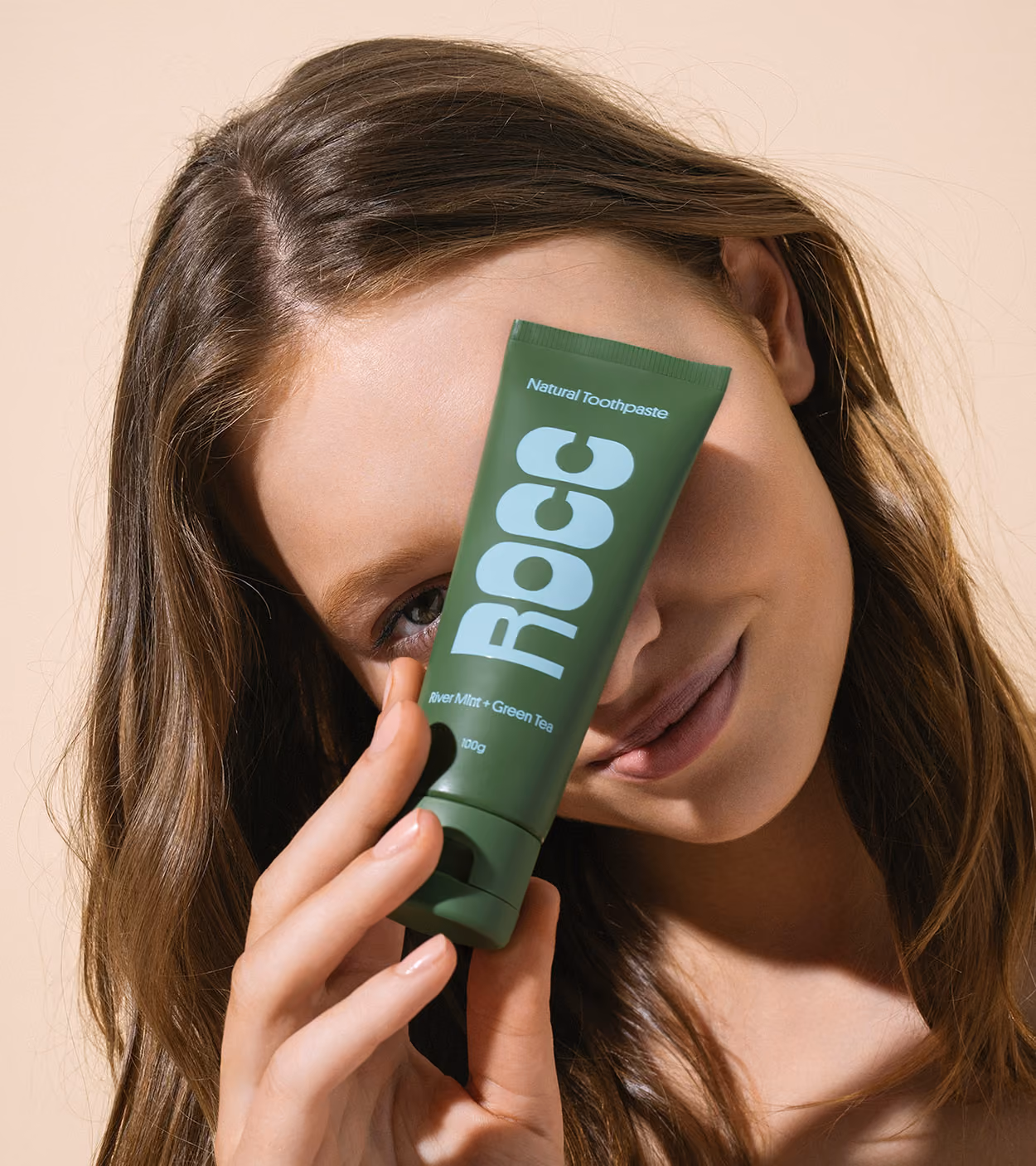 Young woman holding a green tube of Rocc natural toothpaste near her face, smiling softly.