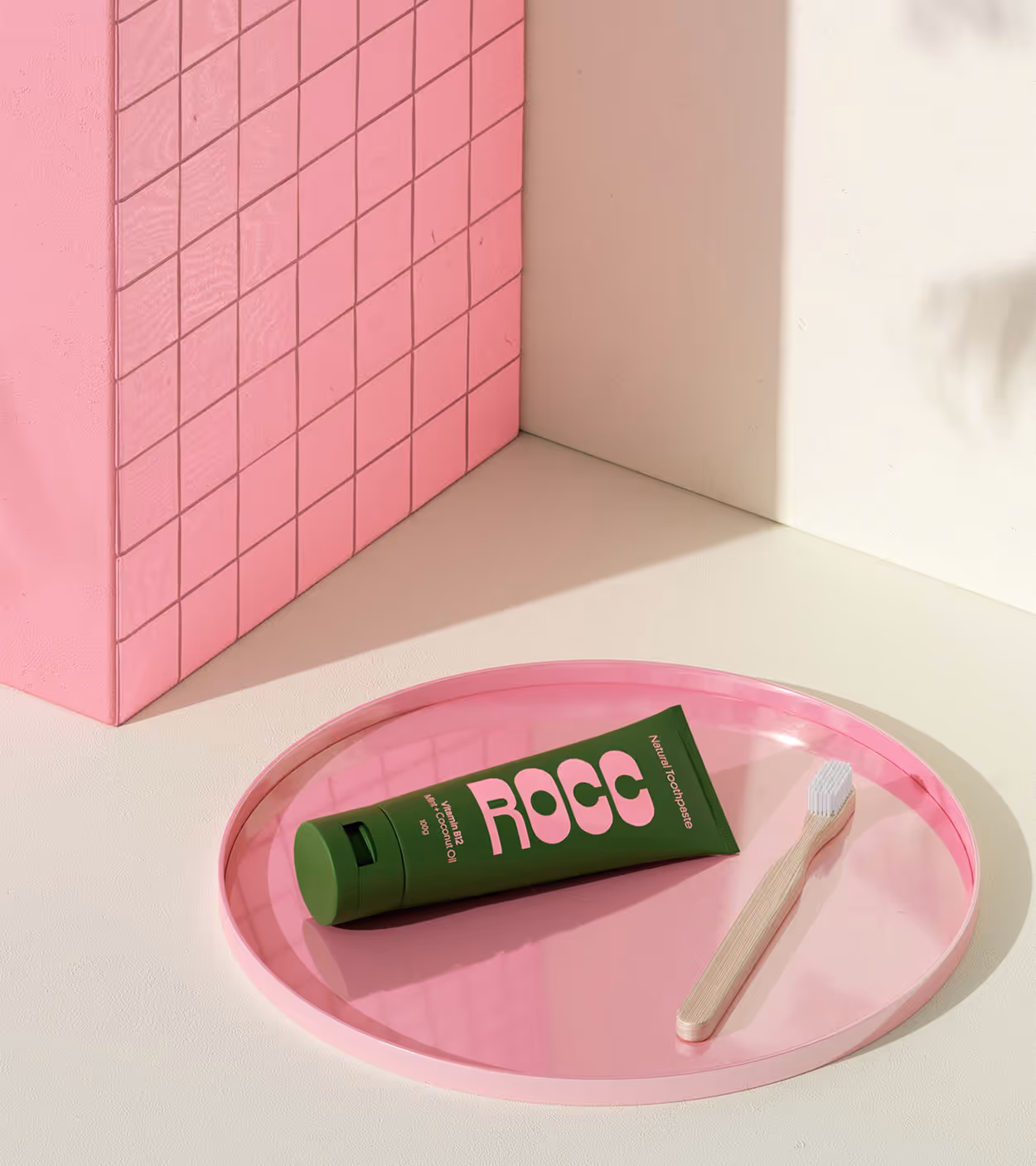 Green tube of ROCC natural toothpaste with a wooden toothbrush on a pink circular tray beside a pink tiled wall.