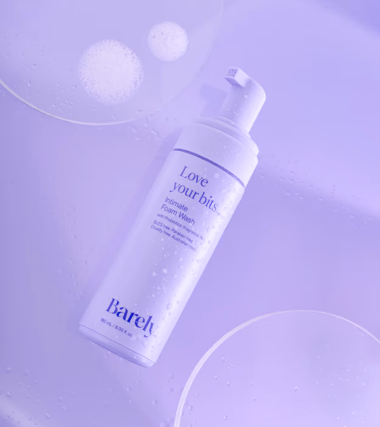 Bottle of Barely intimate foam wash covered in water droplets on a purple background with soap foam bubbles.