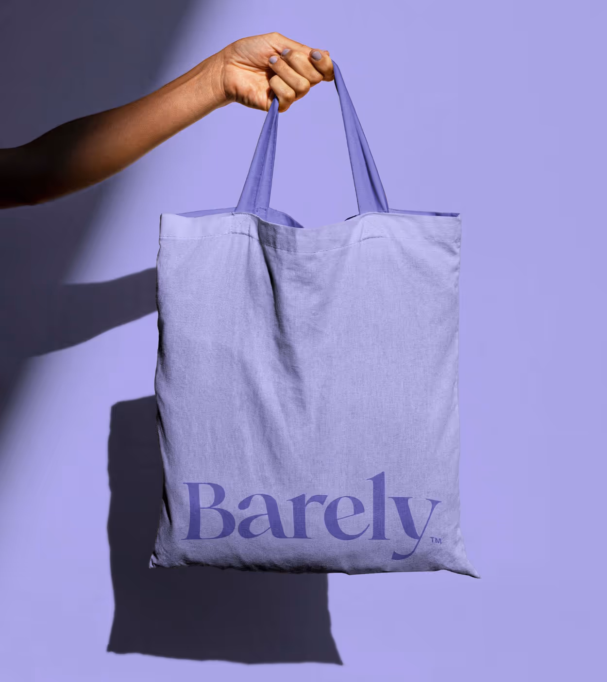 Hand holding a lavender tote bag with the word 'Barely' printed in dark purple on the front against a lavender background.