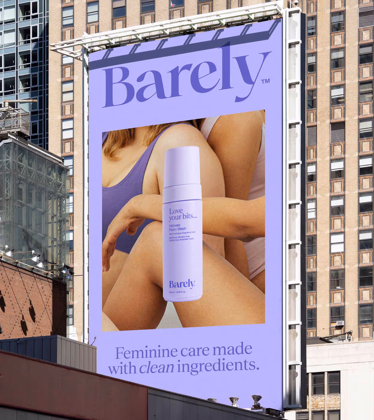 Billboard advertisement for Barely intimate foam wash showing two women embracing and a lavender-colored product bottle with the text 'Love your bits' and 'Feminine care made with clean ingredients.'