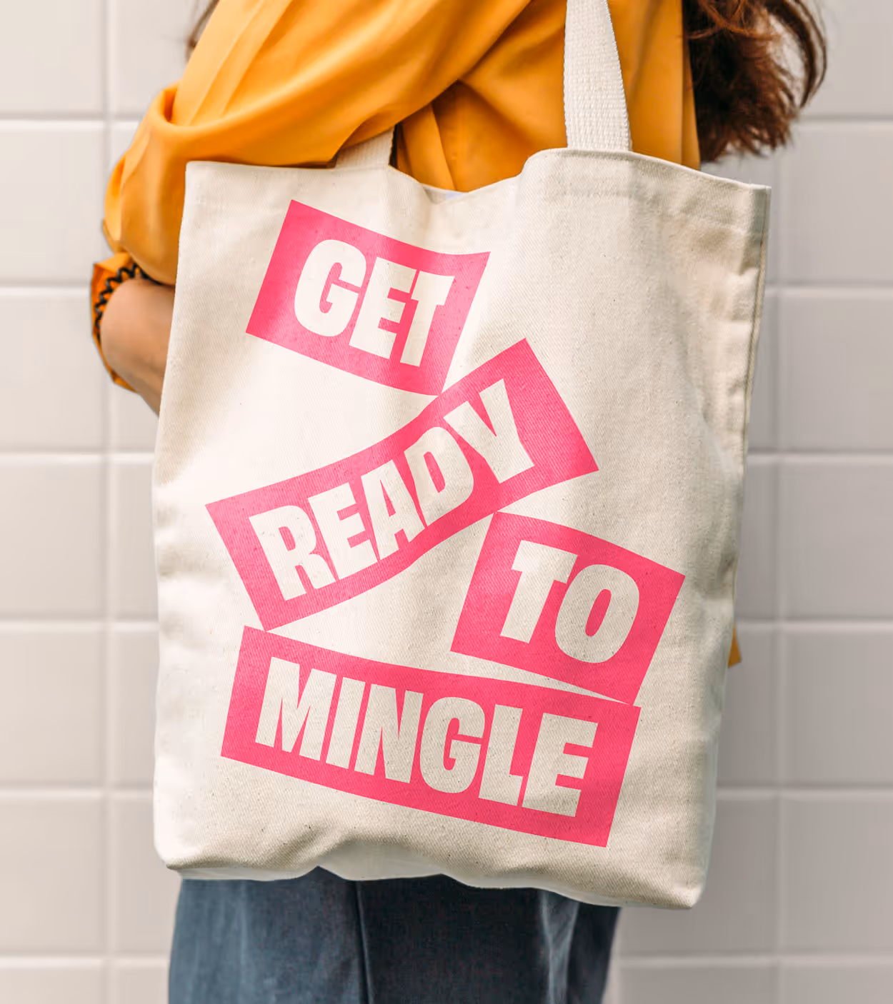 Beige tote bag with bold pink text reading 'GET READY TO MINGLE' carried by a person wearing a mustard yellow top.
