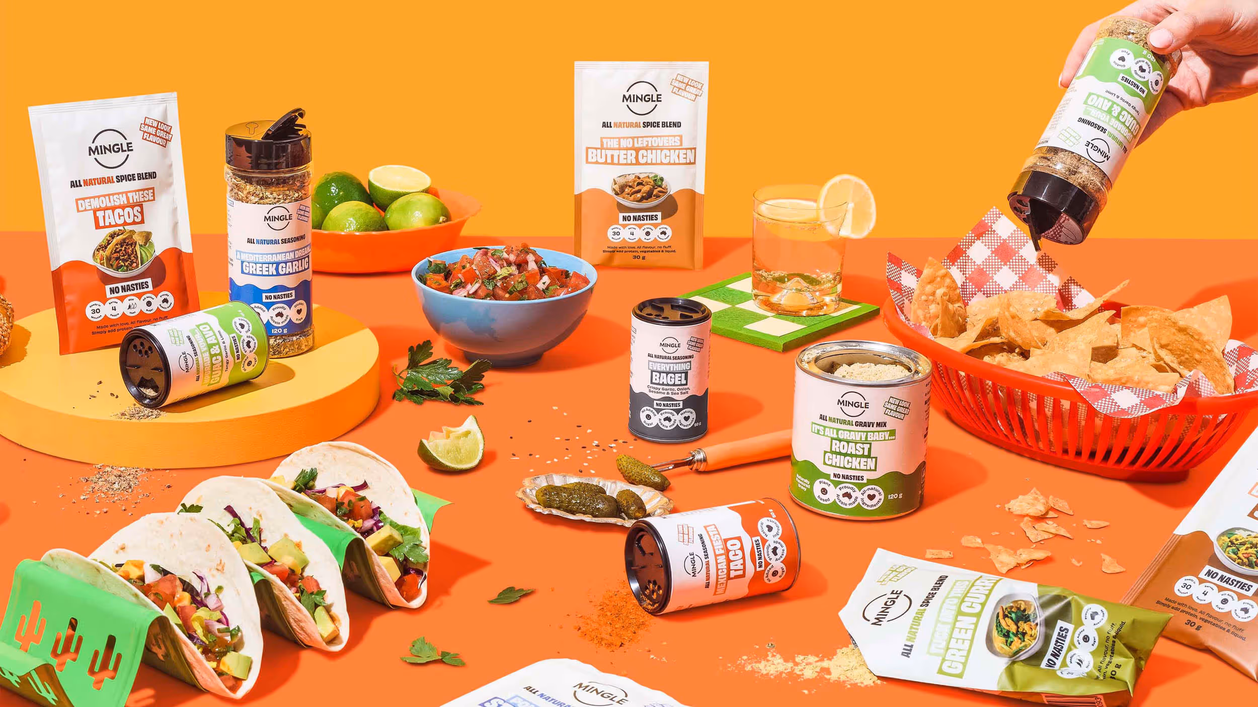 Assortment of Mingle brand spice blends and seasonings arranged with tacos, lemonade with lemon slice, lime fruit, salsa in a blue bowl, pickles, and tortilla chips on an orange background.