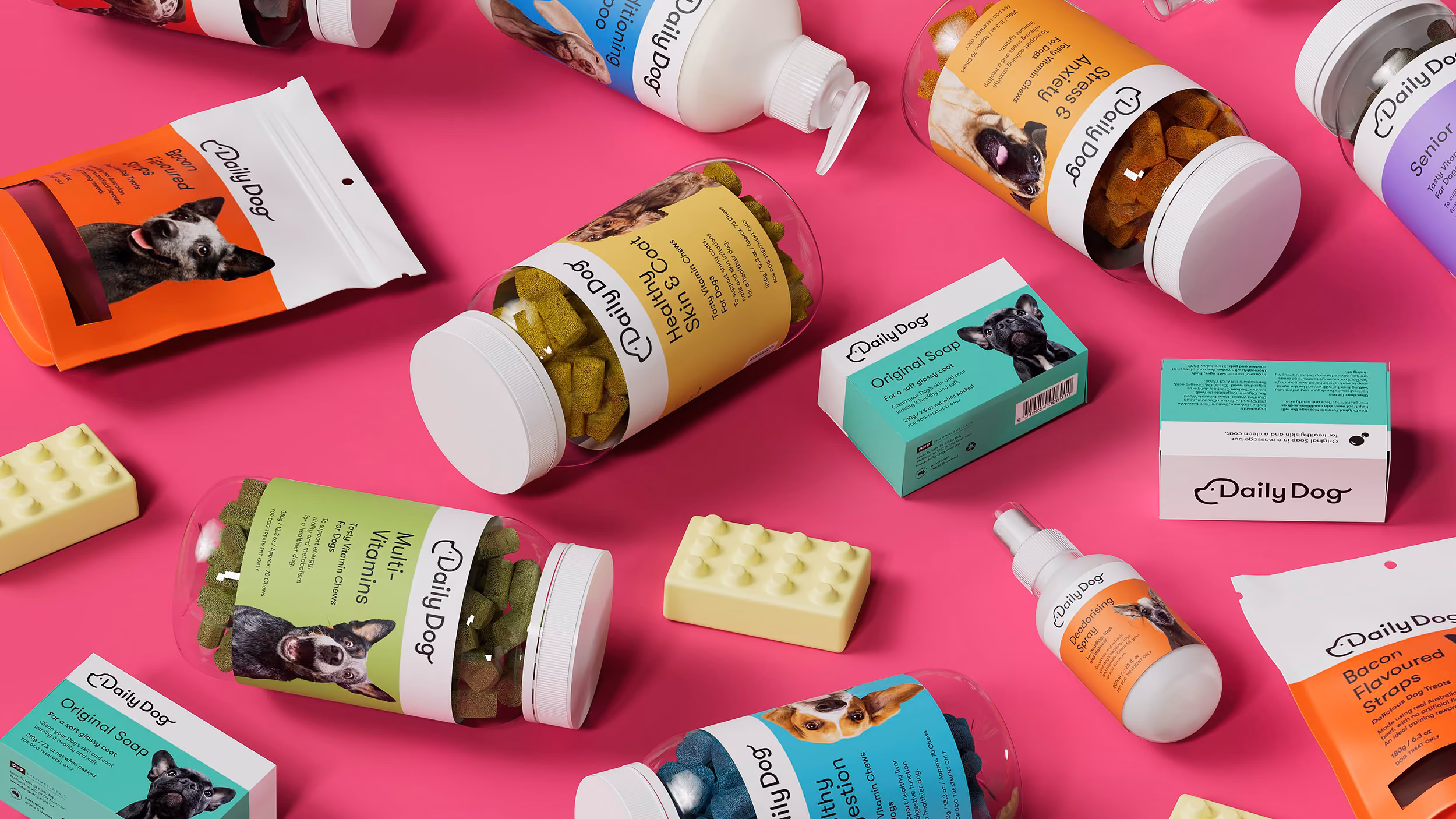 Assorted Daily Dog pet care products including vitamins, conditioning shampoo, original soap, deodorising spray, and flavored treats arranged on a bright pink background.