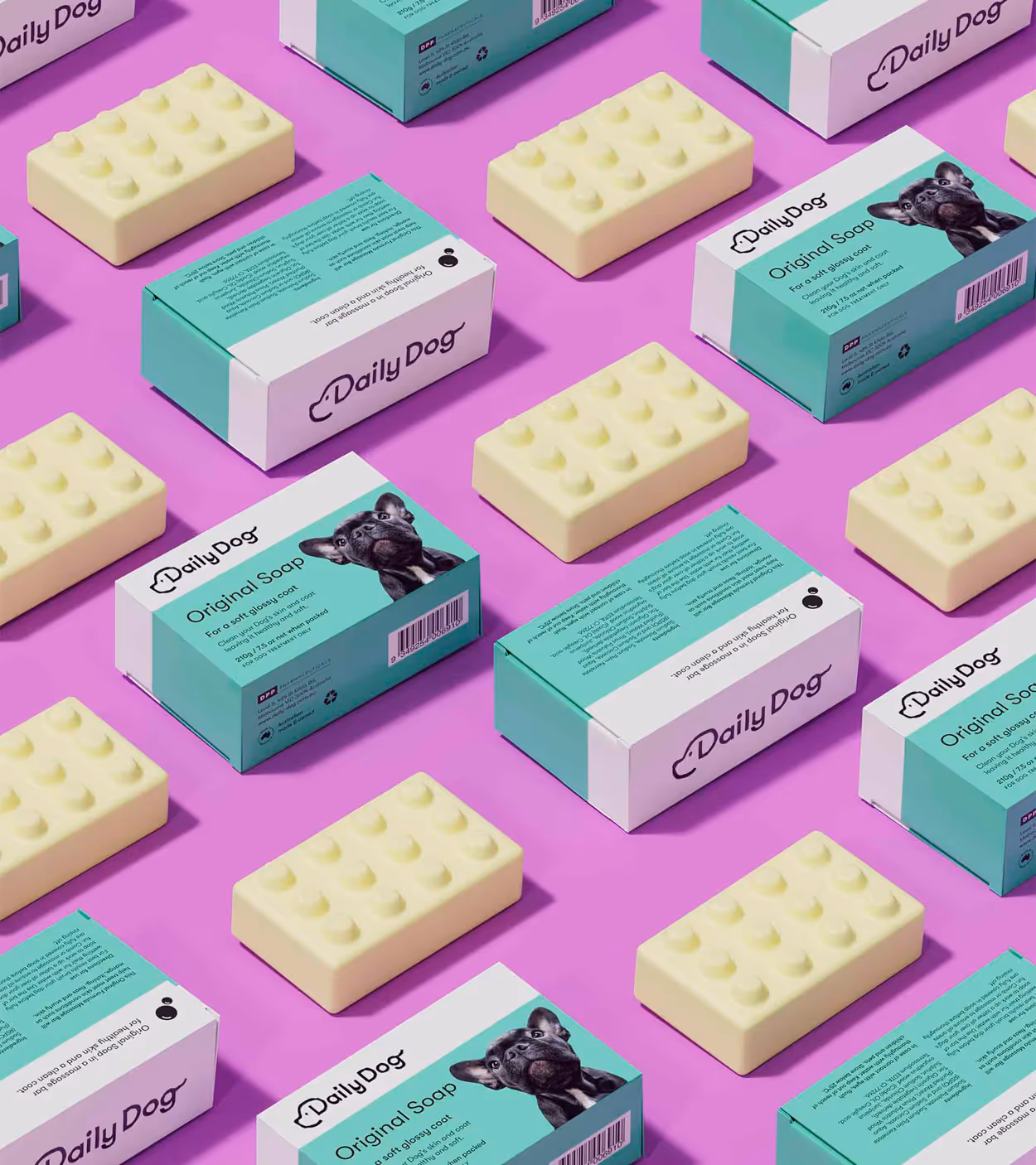 Multiple bars and boxes of Daily Dog Original Soap with a French bulldog image on turquoise and white packaging arranged on a pink background.