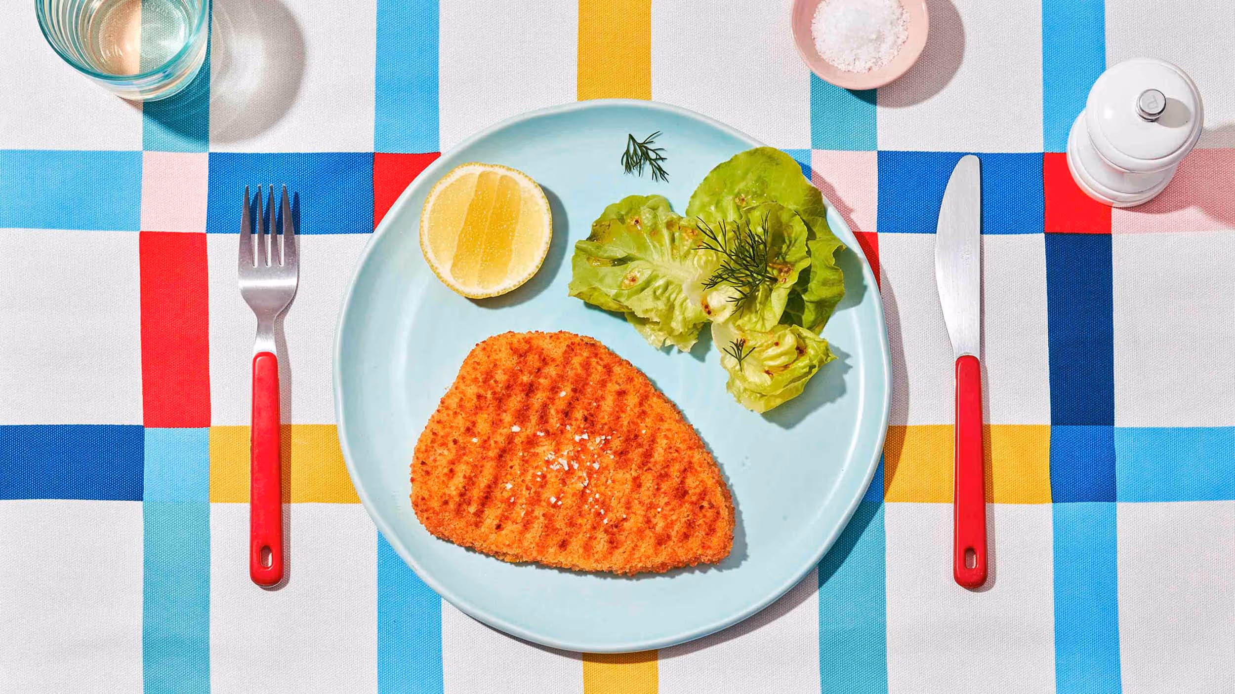 Breaded and grilled chicken cutlet with lettuce, dill, and half a lemon on a light blue plate, set on a colorful checkered tablecloth with red-handled fork and knife.