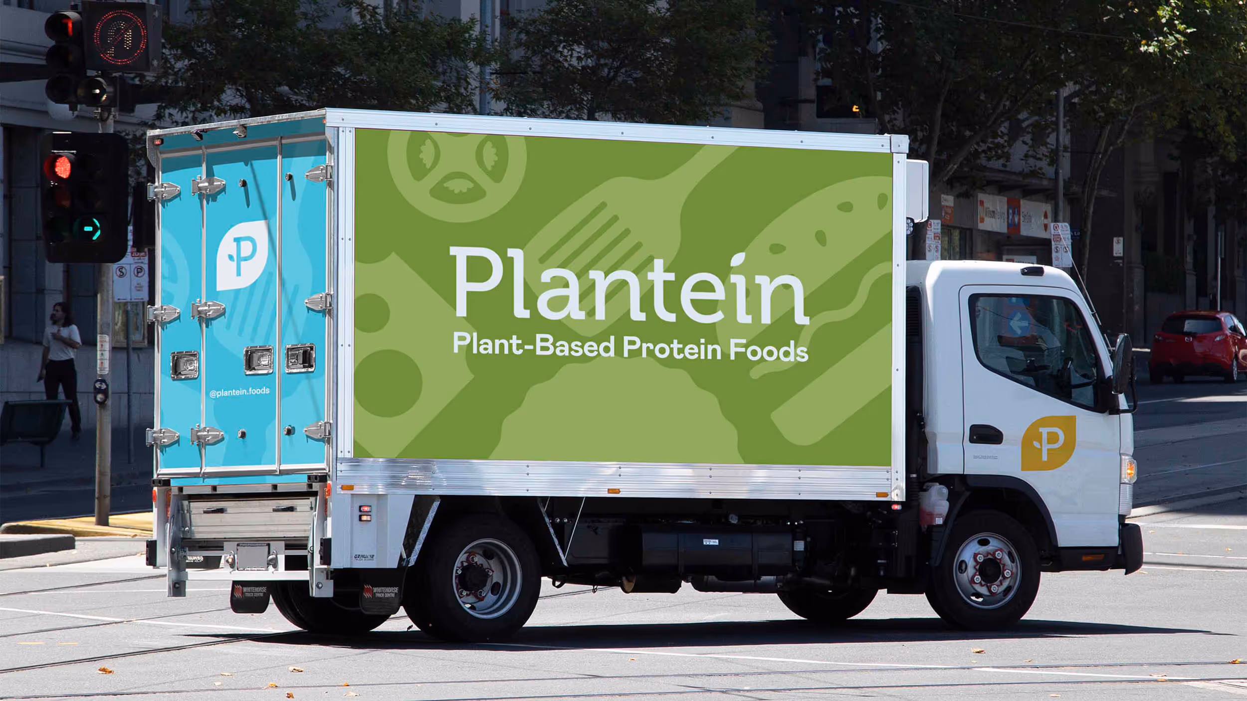 Delivery truck with green and blue panels displaying Plantein logo and text 'Plant-Based Protein Foods' parked at an urban intersection.