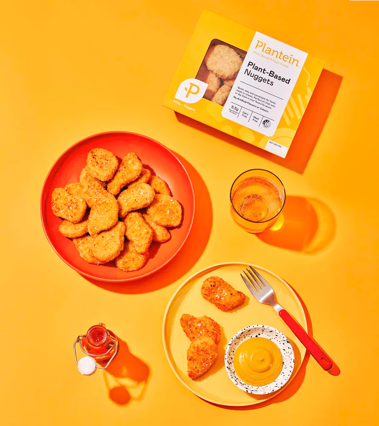 Yellow box of Plantein plant-based nuggets next to a red plate with nuggets, a glass of amber drink, a bottle of hot sauce, and a yellow plate with nuggets, mustard, and a fork.