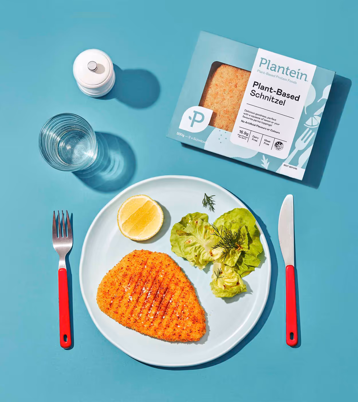Plate with grilled plant-based schnitzel, half a lemon, and lettuce with herbs, surrounded by red-handled fork and knife, clear glass of water, white pepper mill, and plant-based schnitzel packaging on a blue surface.