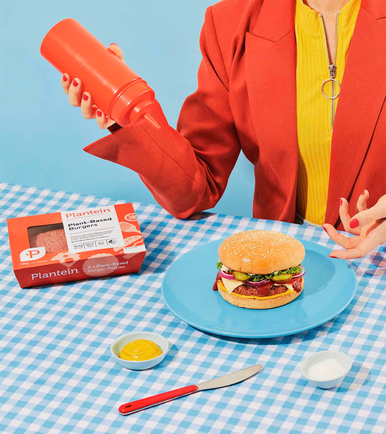 Person wearing red jacket and yellow top holding a red squeeze bottle above a plant-based burger on a blue plate set on a blue and white checkered tablecloth.