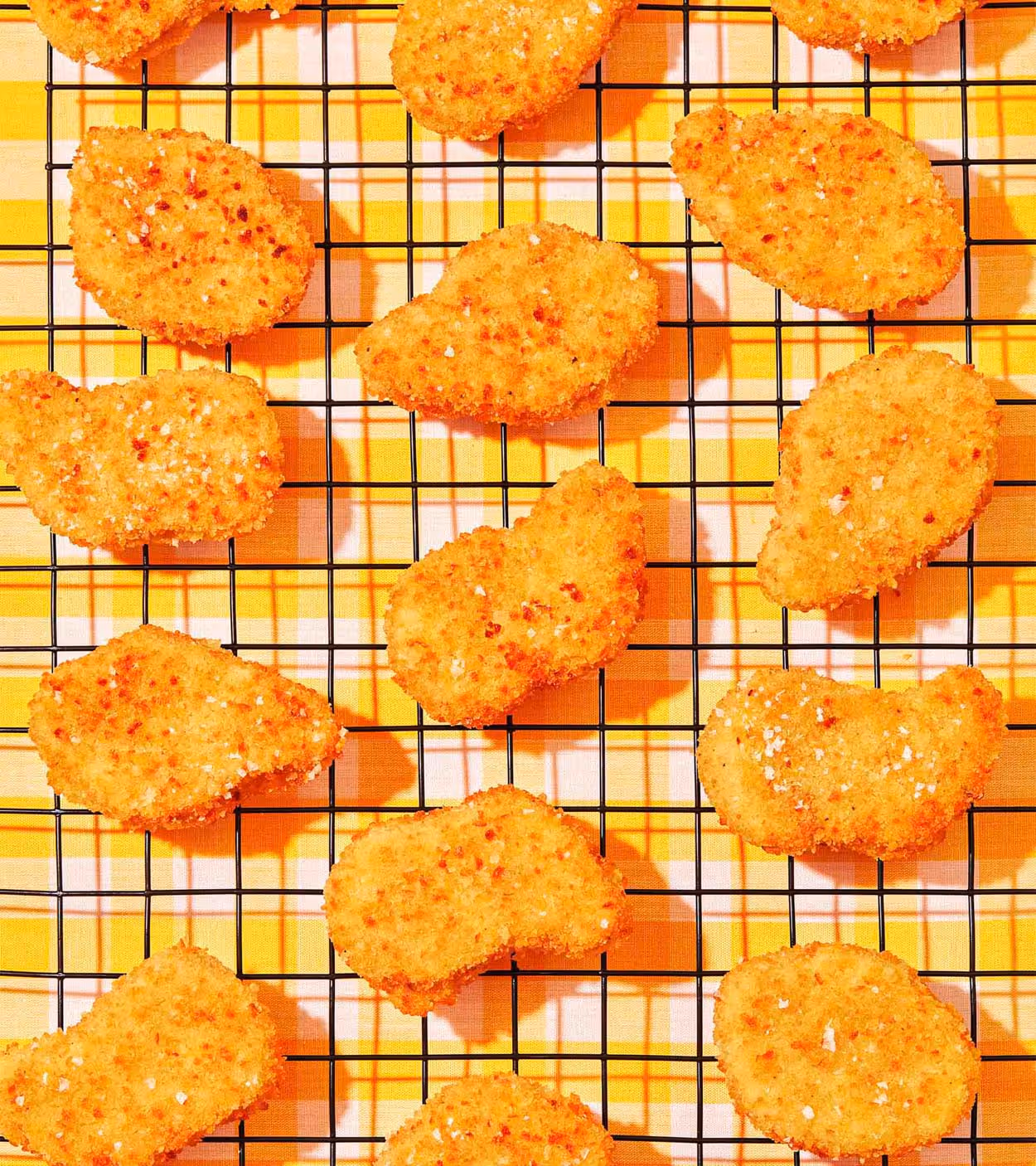 Golden-brown breaded chicken nuggets sprinkled with salt resting on a black cooling rack over a yellow checkered cloth.