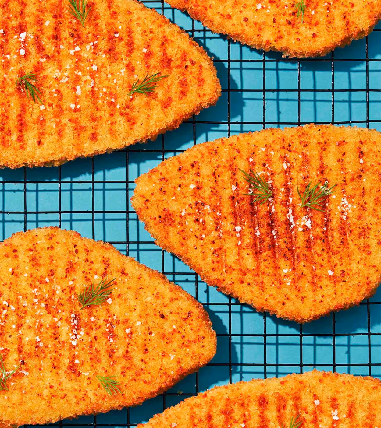 Grilled breaded fish fillets garnished with dill and coarse salt on a black cooling rack over a blue surface.