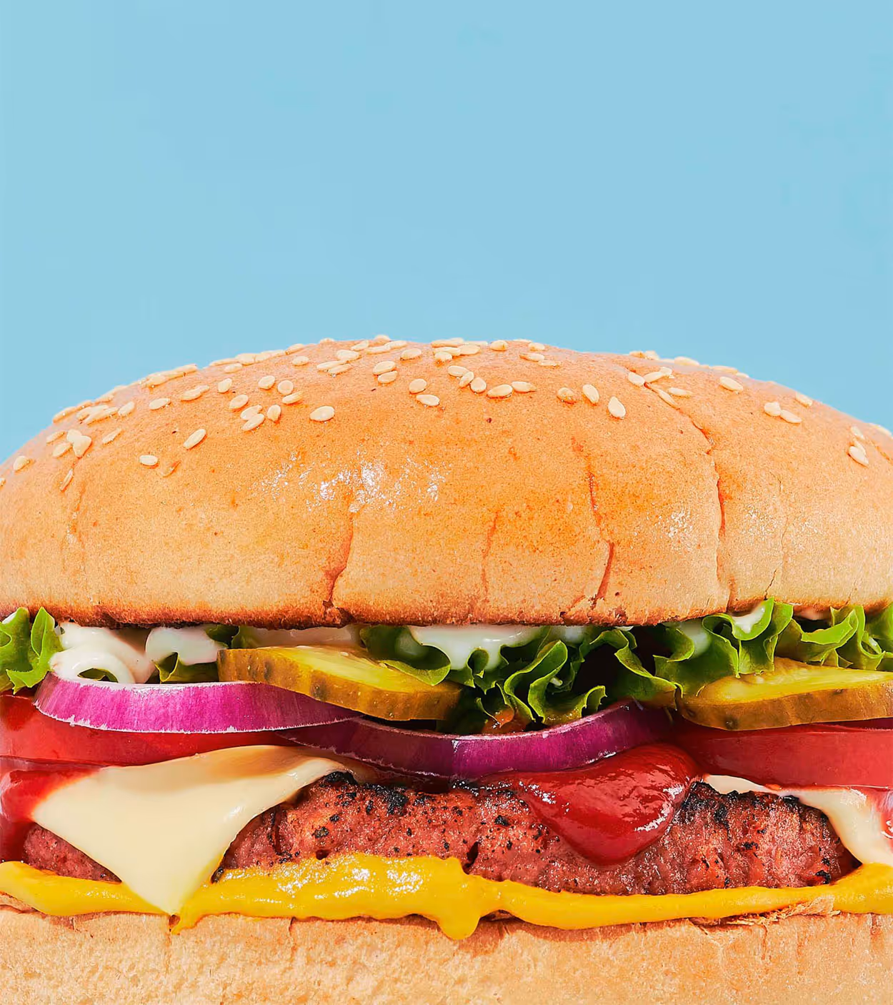 Close-up of a cheeseburger with lettuce, pickles, red onion, tomato, ketchup, mustard, and sesame seed bun against a blue background.