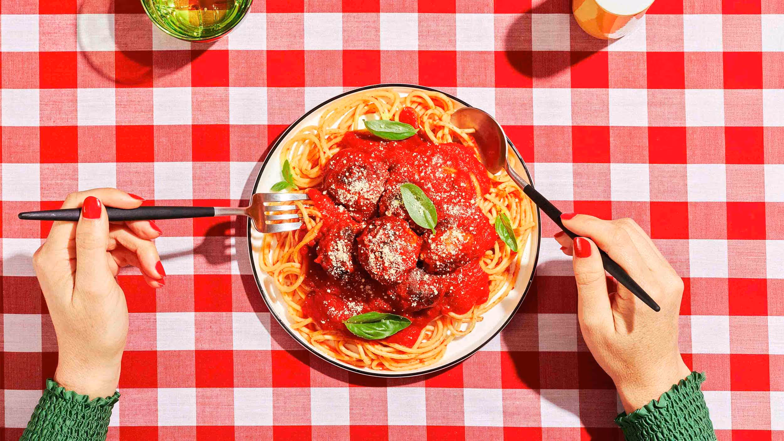 Plate of spaghetti with meatballs and tomato sauce garnished with basil leaves on a red and white checkered tablecloth, with hands holding a fork and spoon.