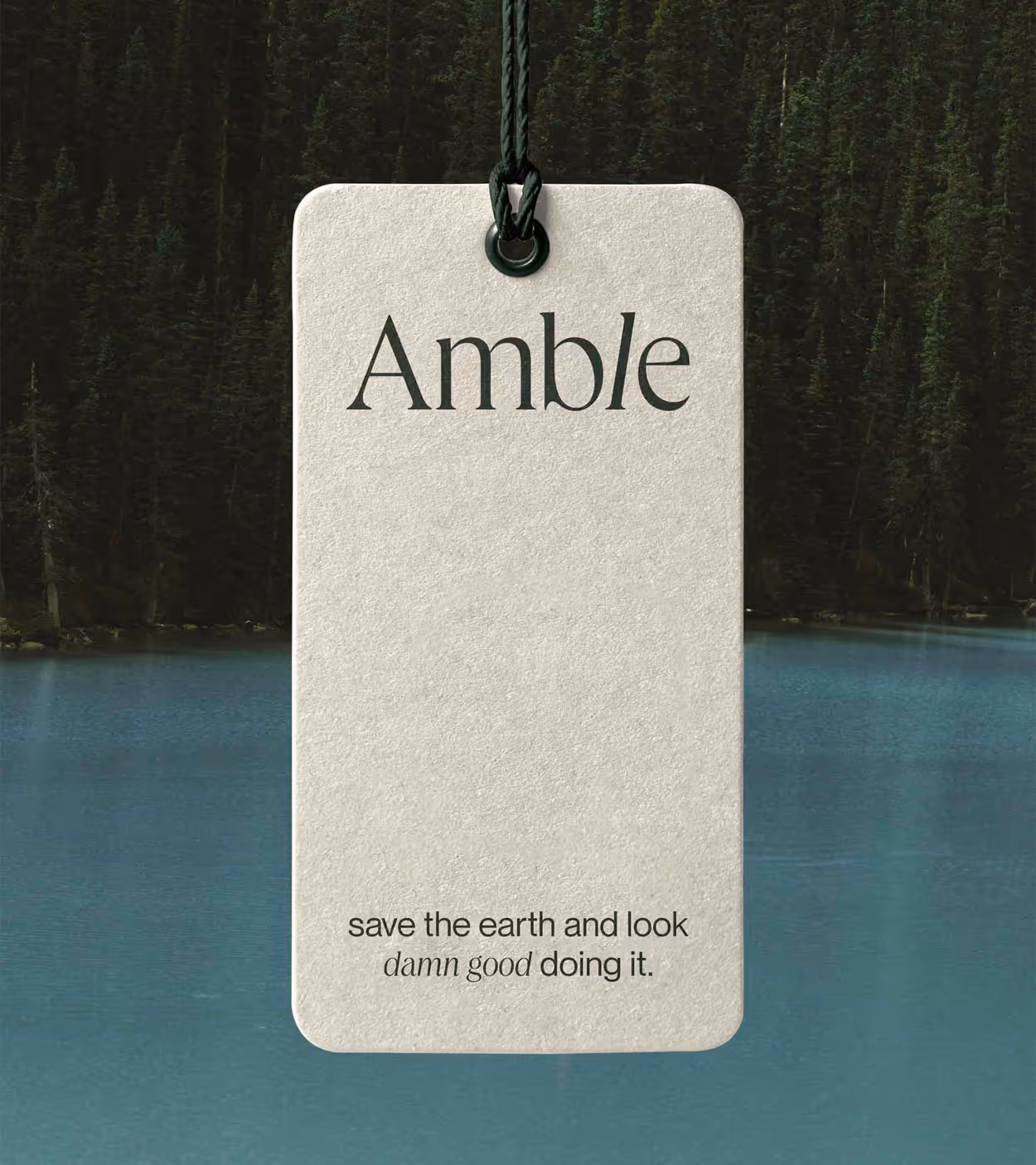 Beige rectangular tag hanging by a dark string over a lake with forest background, with text 'Amble' and 'save the earth and look damn good doing it.'