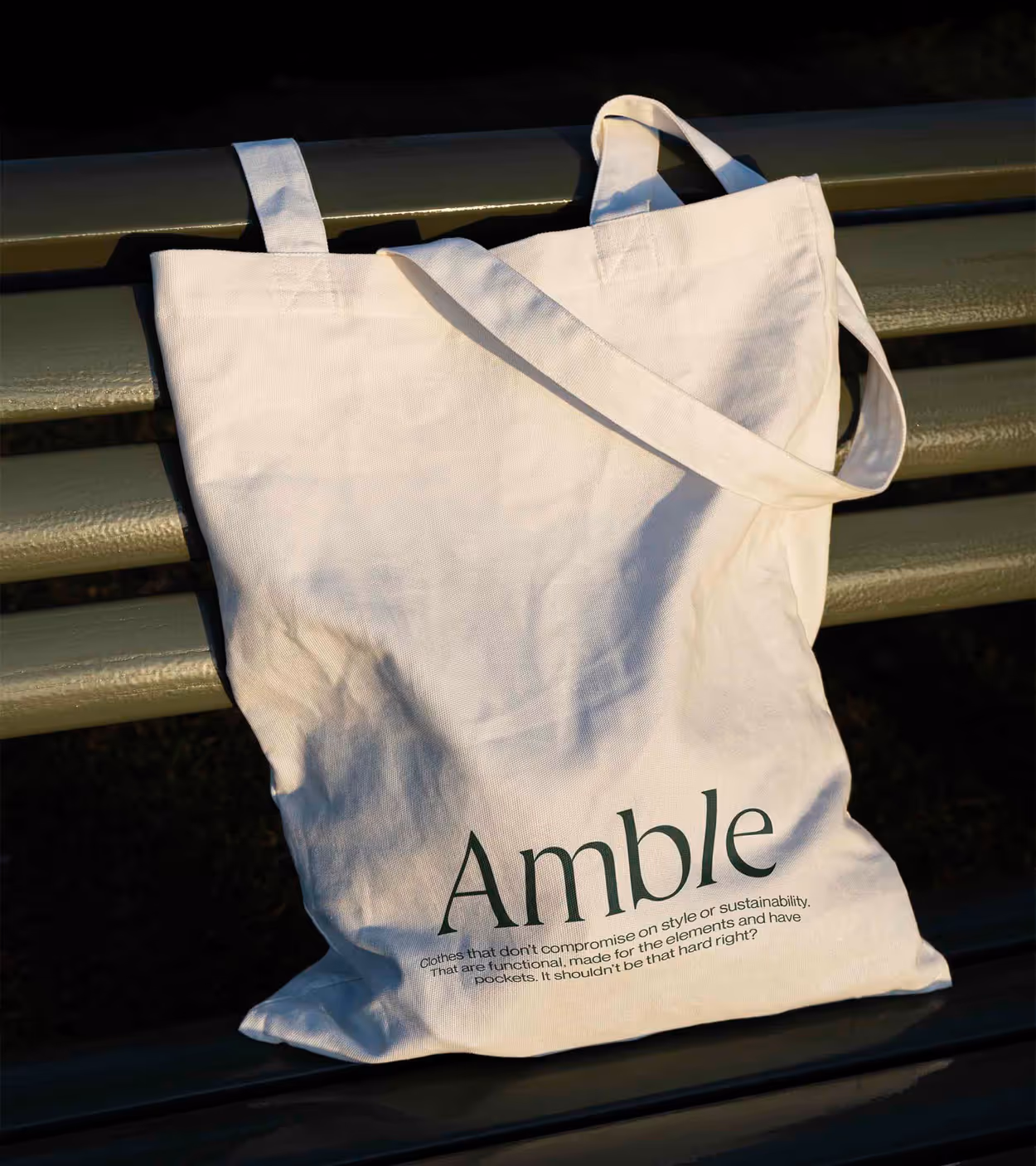 White canvas tote bag with the word 'Amble' and a sustainability message, resting on an olive green park bench.