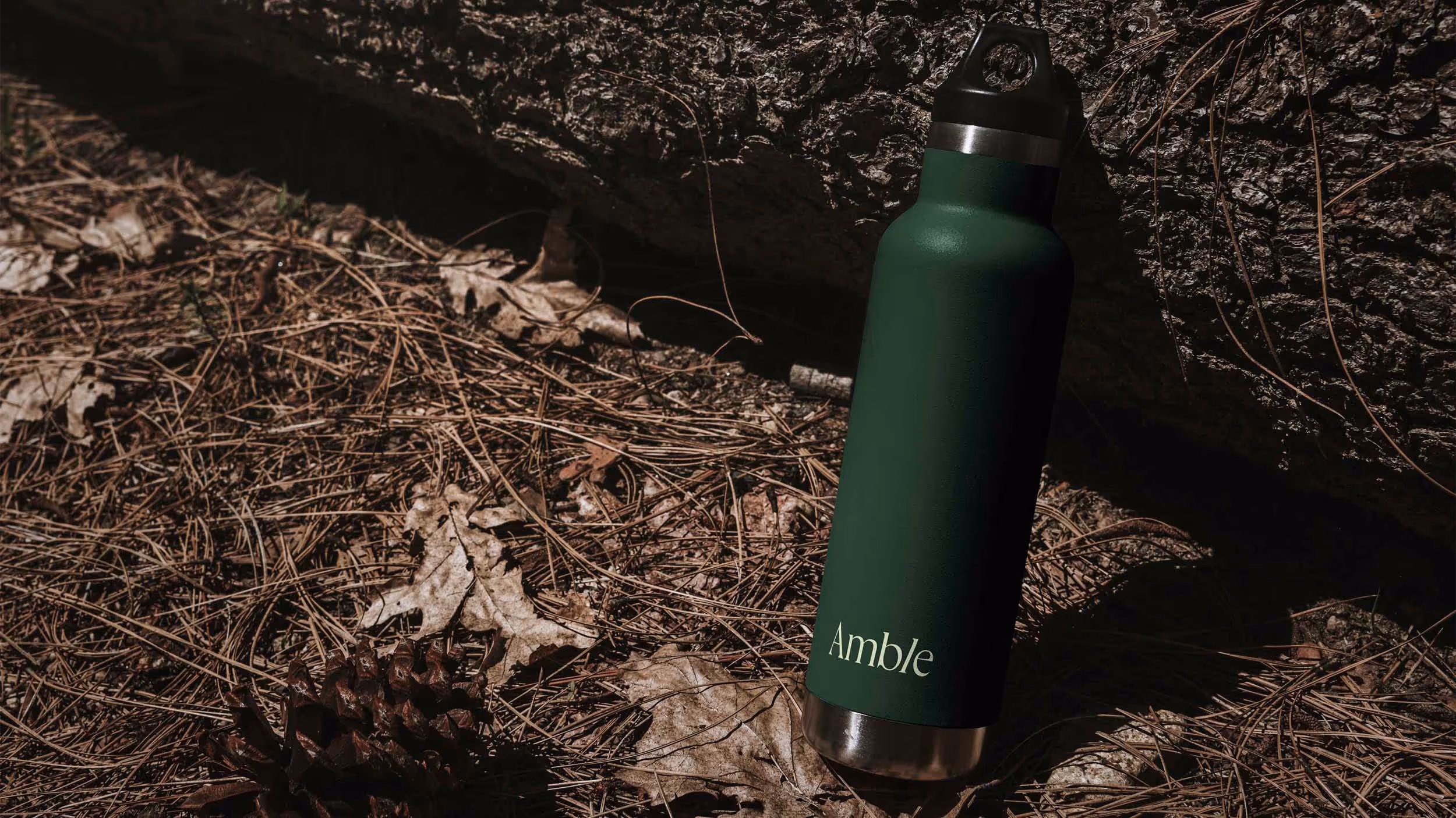 Dark green insulated water bottle with black cap and ‘Amble’ logo placed on pine needles and dry leaves near a textured rock.
