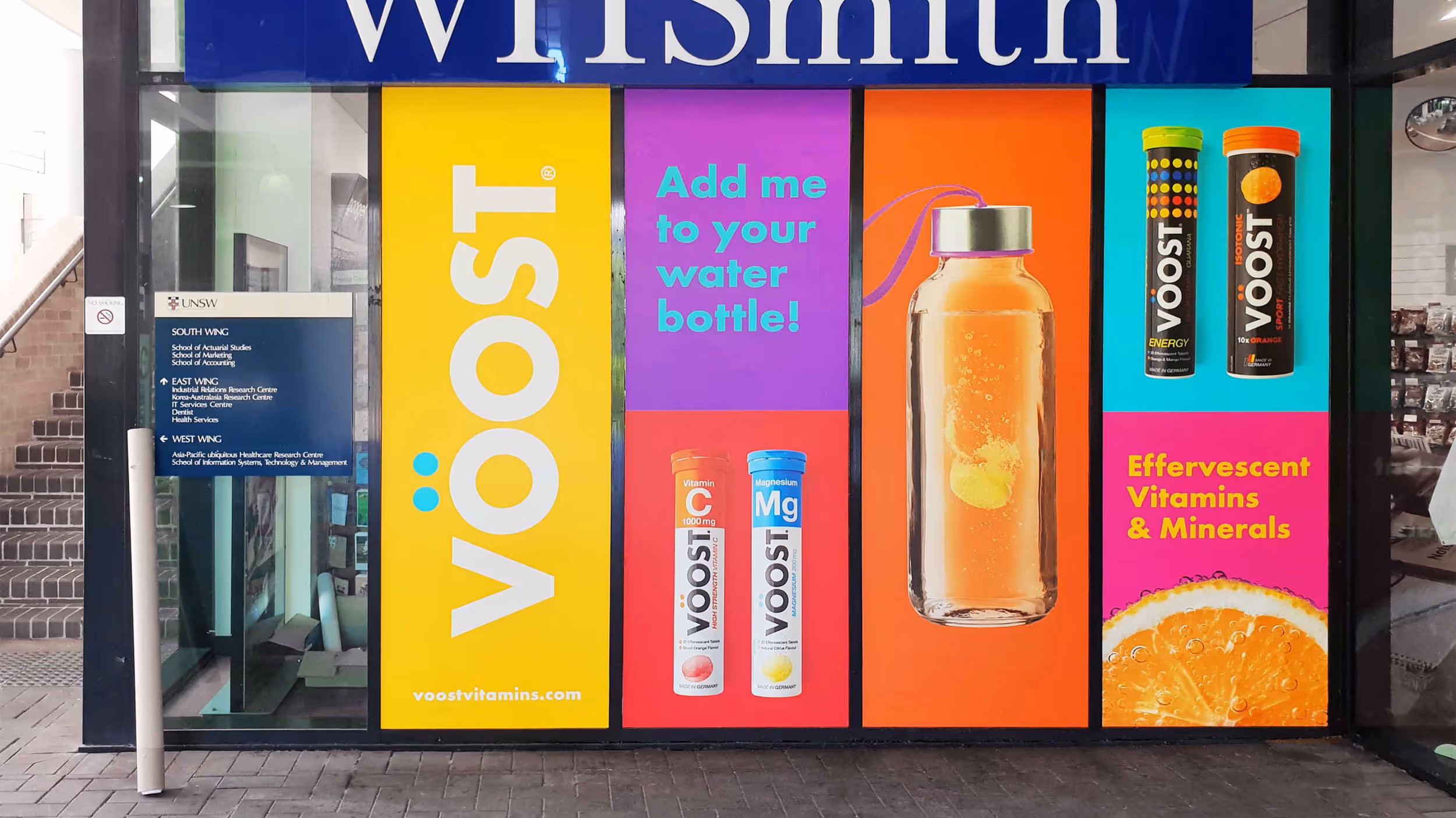 Storefront window at WHSmith displaying colorful ads for Voost effervescent vitamins with text 'Add me to your water bottle!' and images of vitamin tubes and a water bottle.
