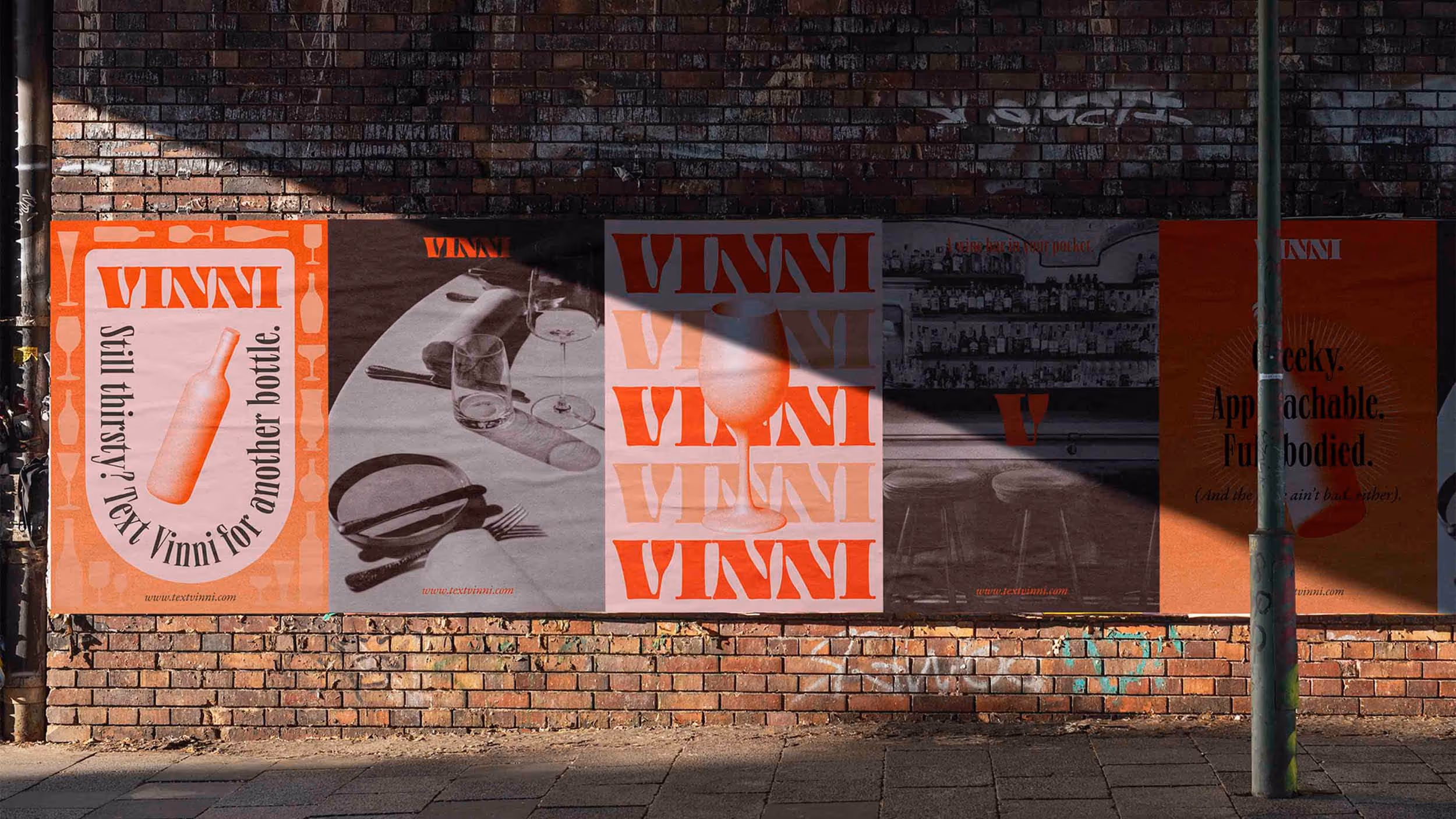 Brick wall with five orange and pink street posters advertising VINNI wine, featuring text, wine bottles, glasses, and bar scenes, partially shaded by a lamp post.