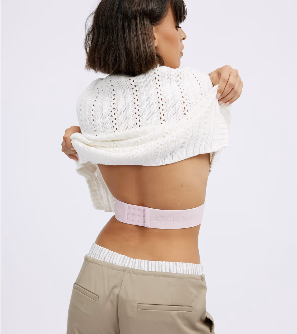 Woman lifting white knitted sweater showing back and light pink bra band with 'BASIC BAN' text.