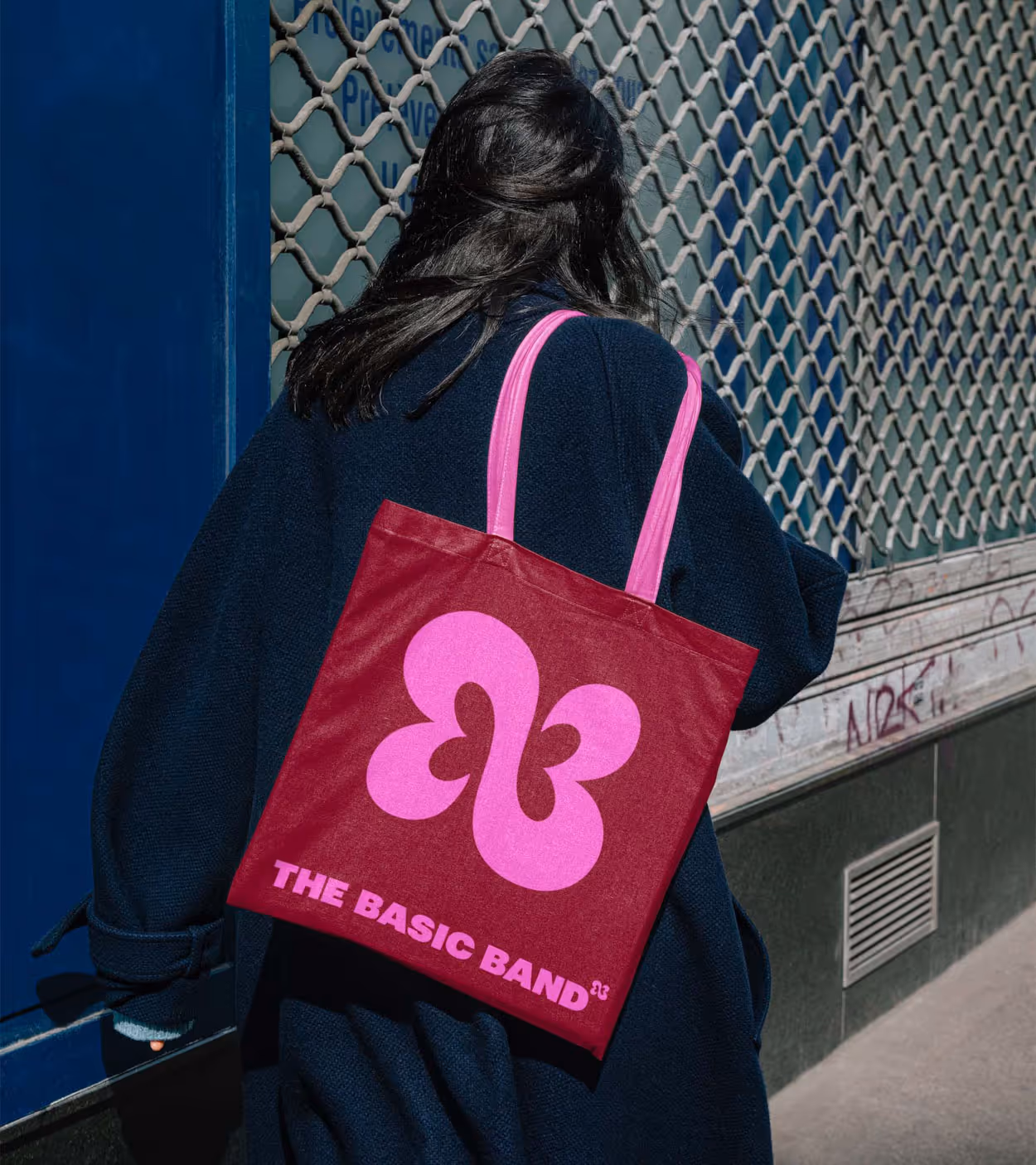 Person with long dark hair wearing a dark coat carrying a red tote bag with pink handles and logo, reading 'THE BASIC BAND'.