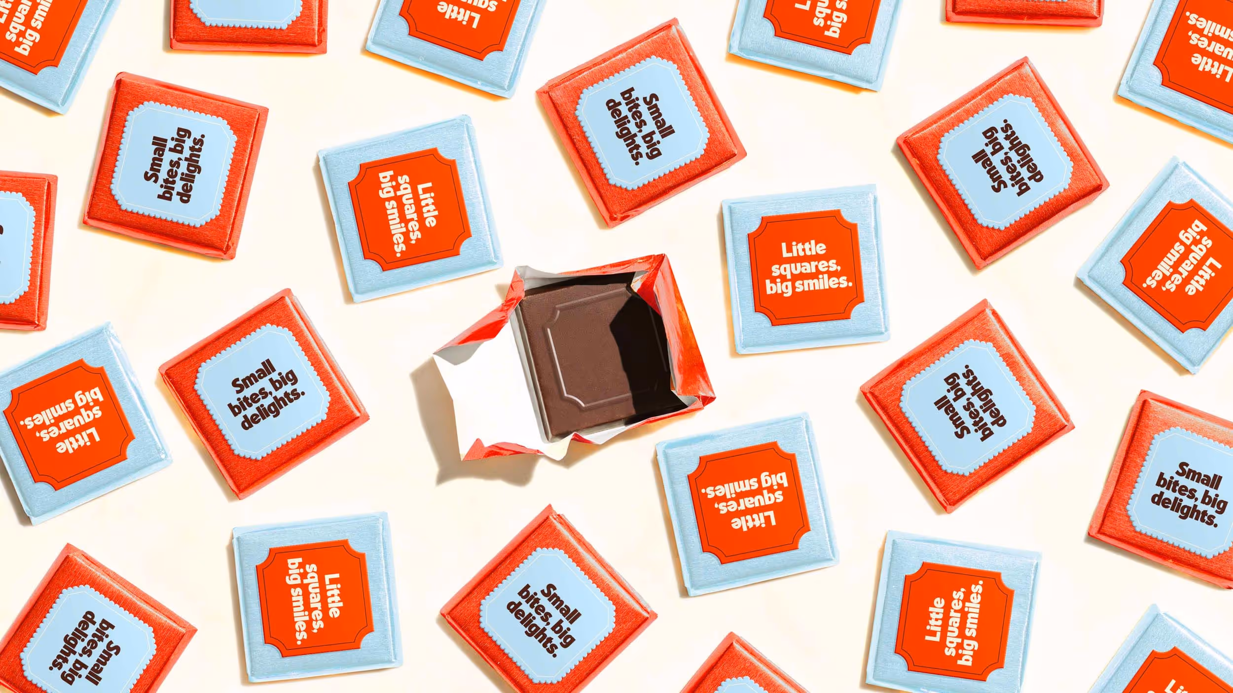 Assorted small chocolate squares in red and blue wrappers with phrases like 'Small bites, big delights' and 'Little squares, big smiles', one unwrapped square in the center.