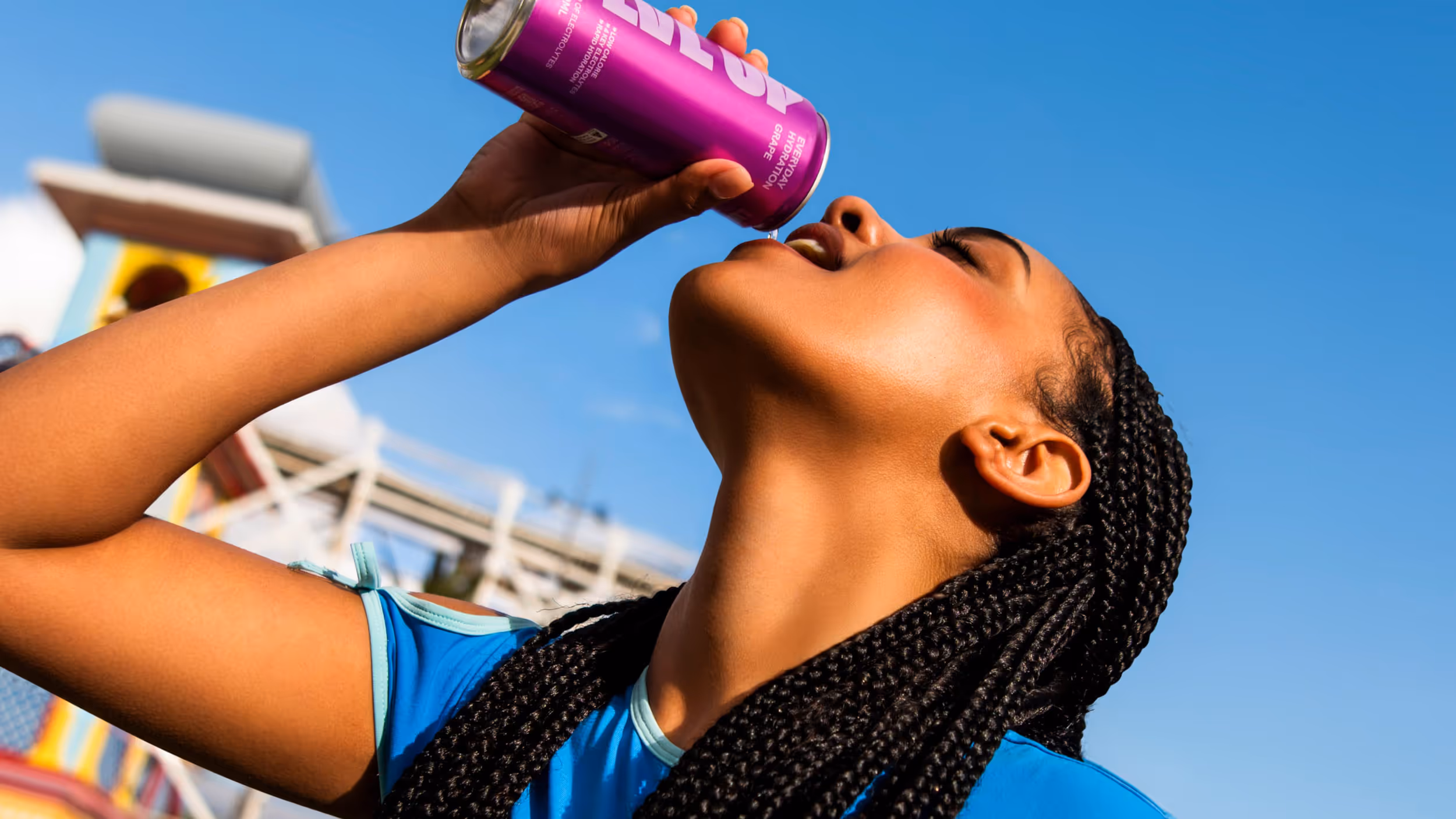 Woman with braided hair wearing a blue top drinking from a purple can labeled 'everyday hydration grape' against a clear blue sky.