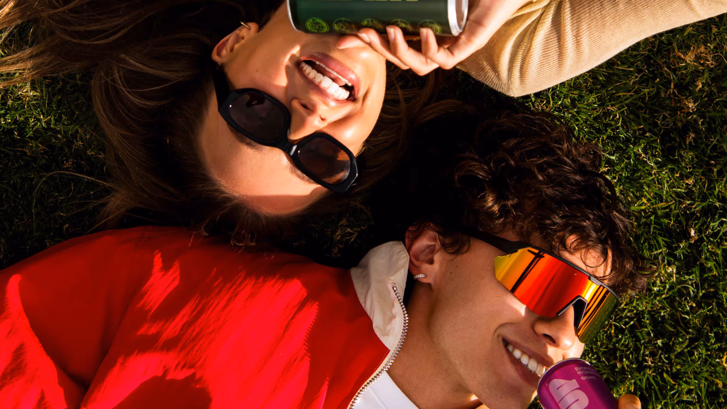 Smiling man and woman lying on grass wearing sunglasses, each drinking from a can.