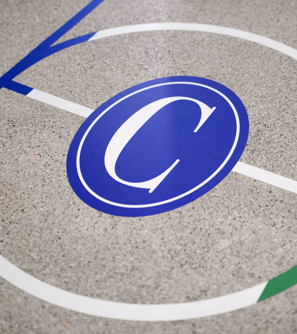 Blue circular logo with a white letter C inside, painted on a textured gray concrete surface with white and blue court lines intersecting near it.