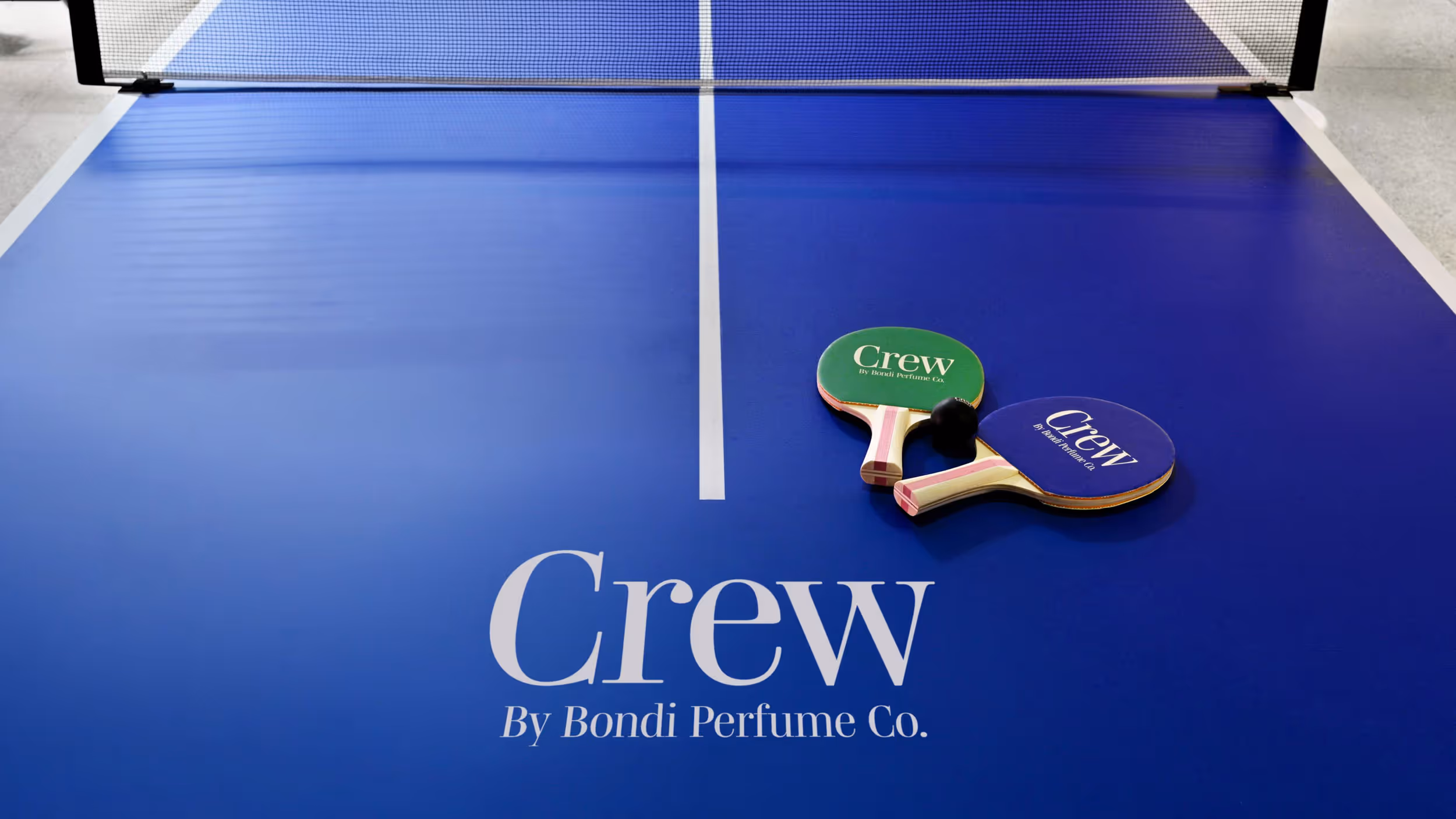 Blue ping pong table with two paddles and a ball, featuring the text 'Crew By Bondi Perfume Co.' on the table surface.