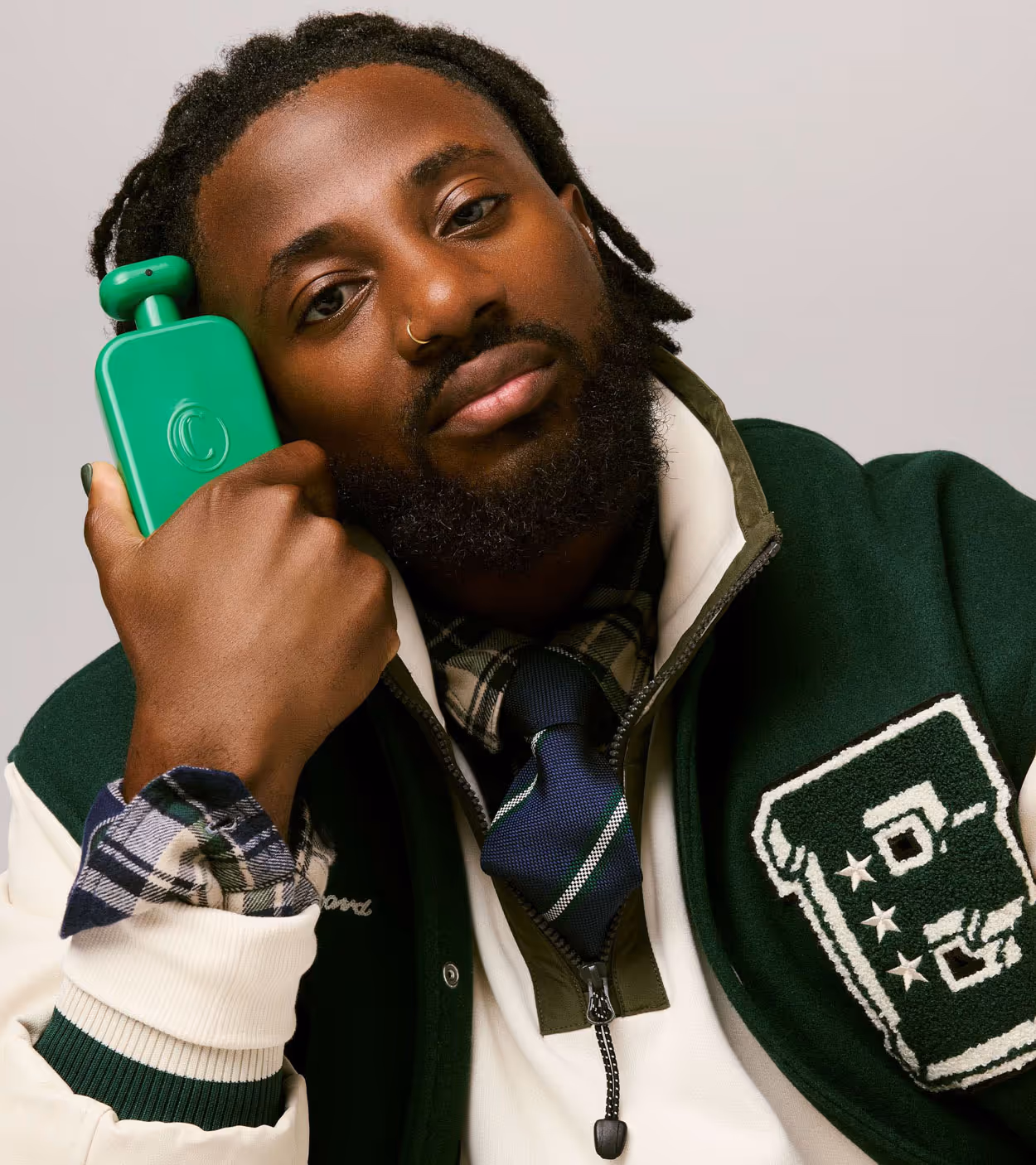 Bearded man with nose ring wearing a green and white varsity jacket holding a green hot water bottle to his head.