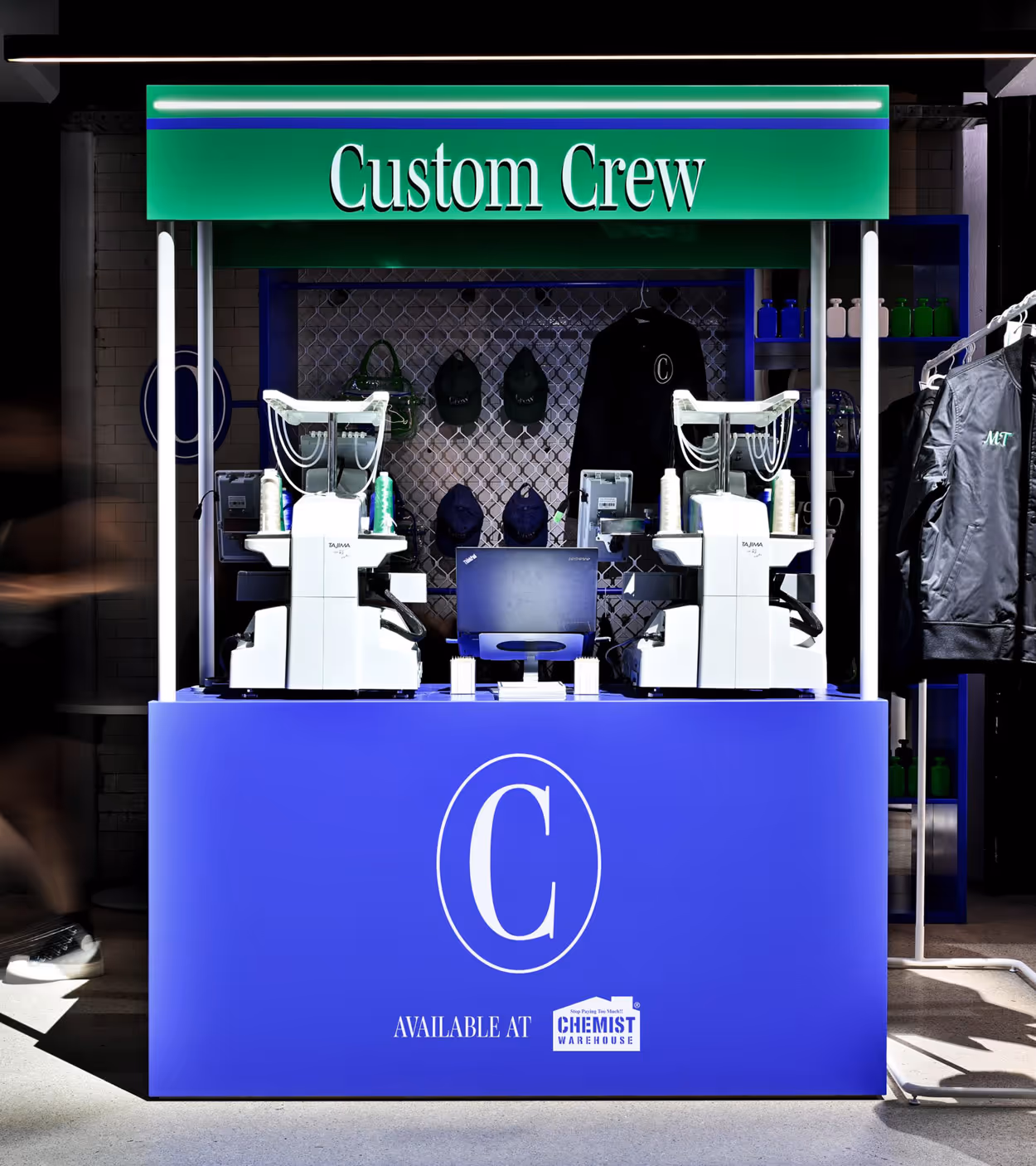Custom Crew embroidery station with two sewing machines and computer on a blue counter branded with a large C logo and Chemist Warehouse availability.