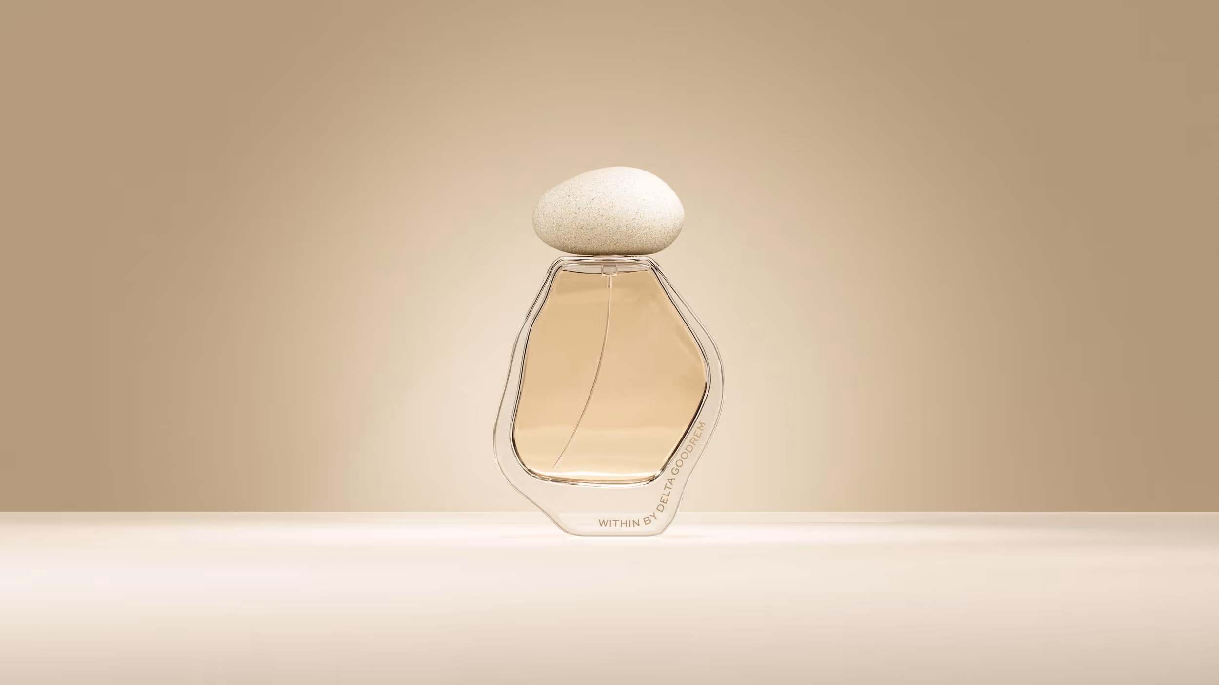 Transparent perfume bottle with beige liquid and a rounded stone cap on a light beige surface.