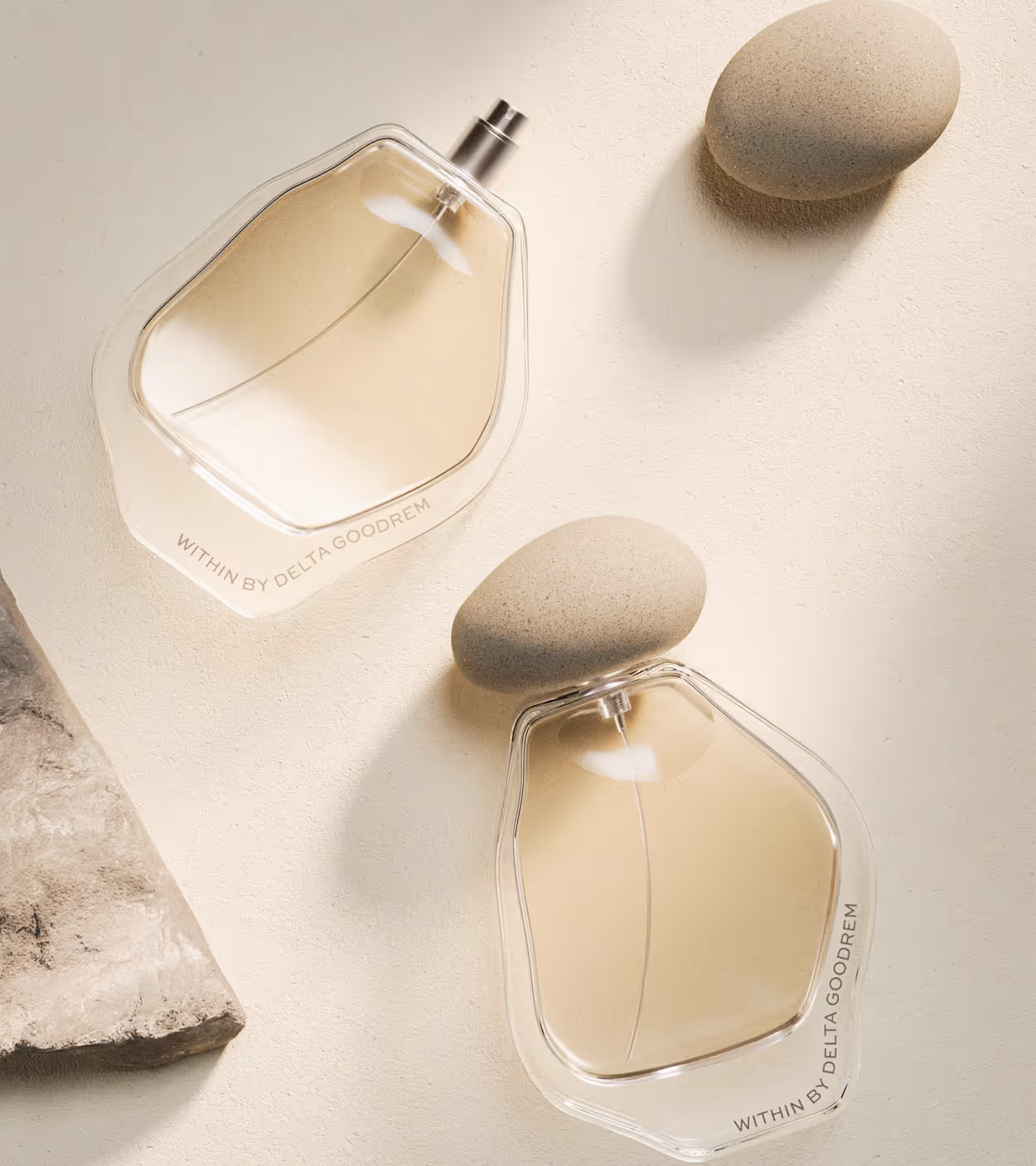 Two clear perfume bottles with beige liquid labeled 'Within by Delta Goodrem' next to two smooth beige stones on a textured cream surface.