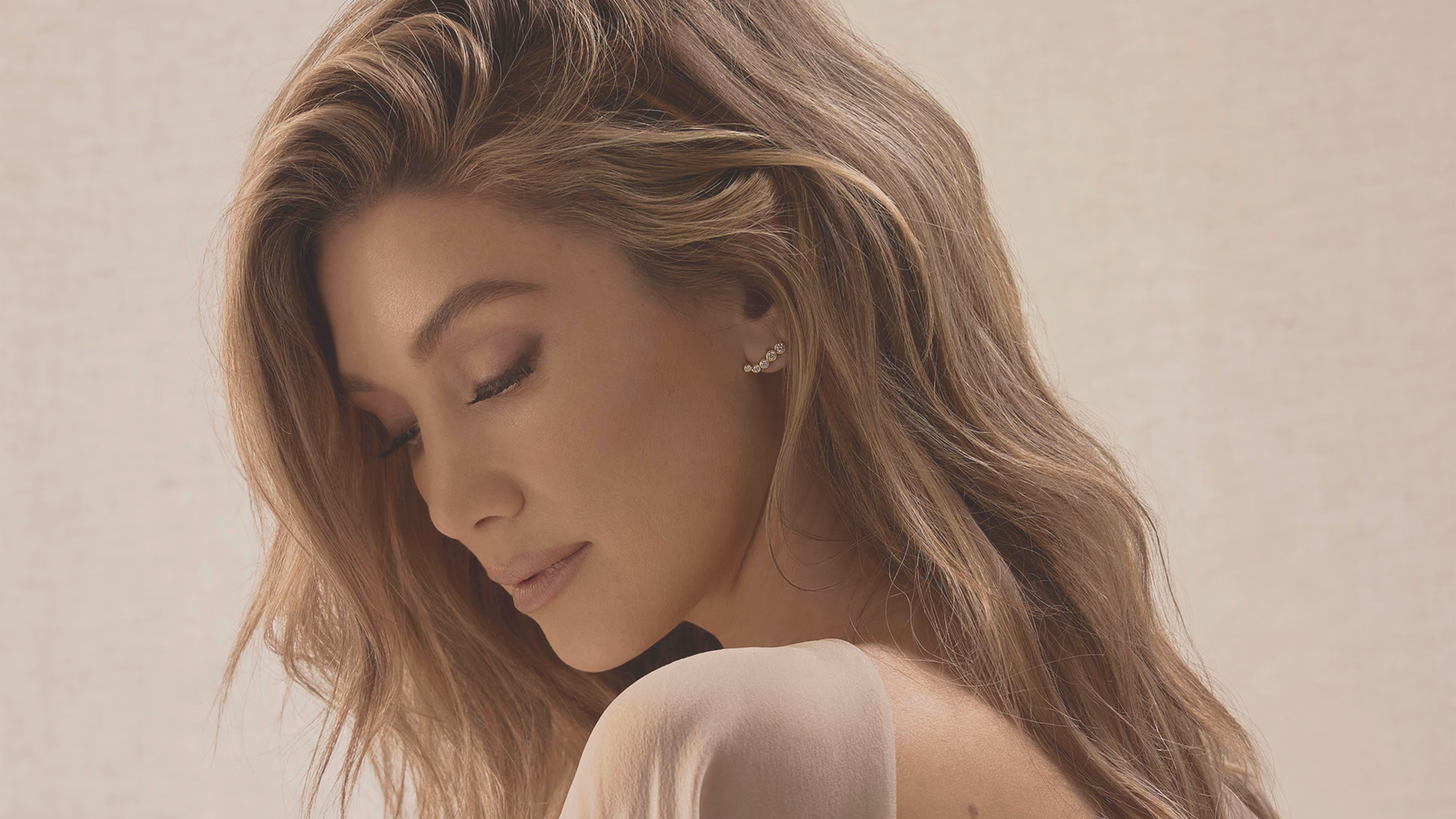 Woman with long wavy light brown hair and closed eyes wearing a white top and diamond ear climber earrings.