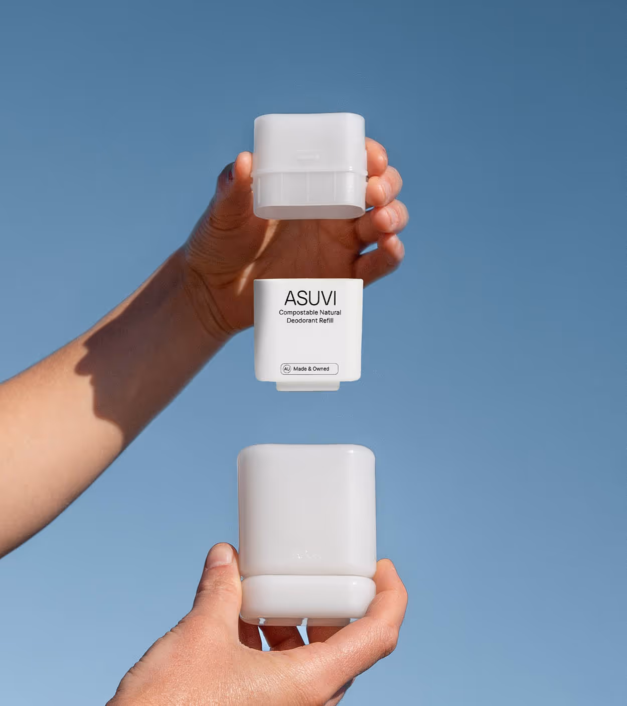 Two hands holding a white ASUVI compostable natural deodorant refill container against a clear blue sky.