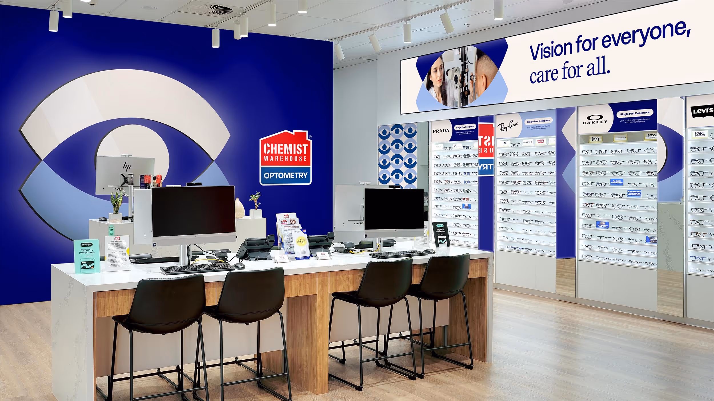 Interior of a Chemist Warehouse Optometry store with computer desks, black chairs, and eyeglass displays featuring brands like Ray-Ban, Oakley, and Prada.