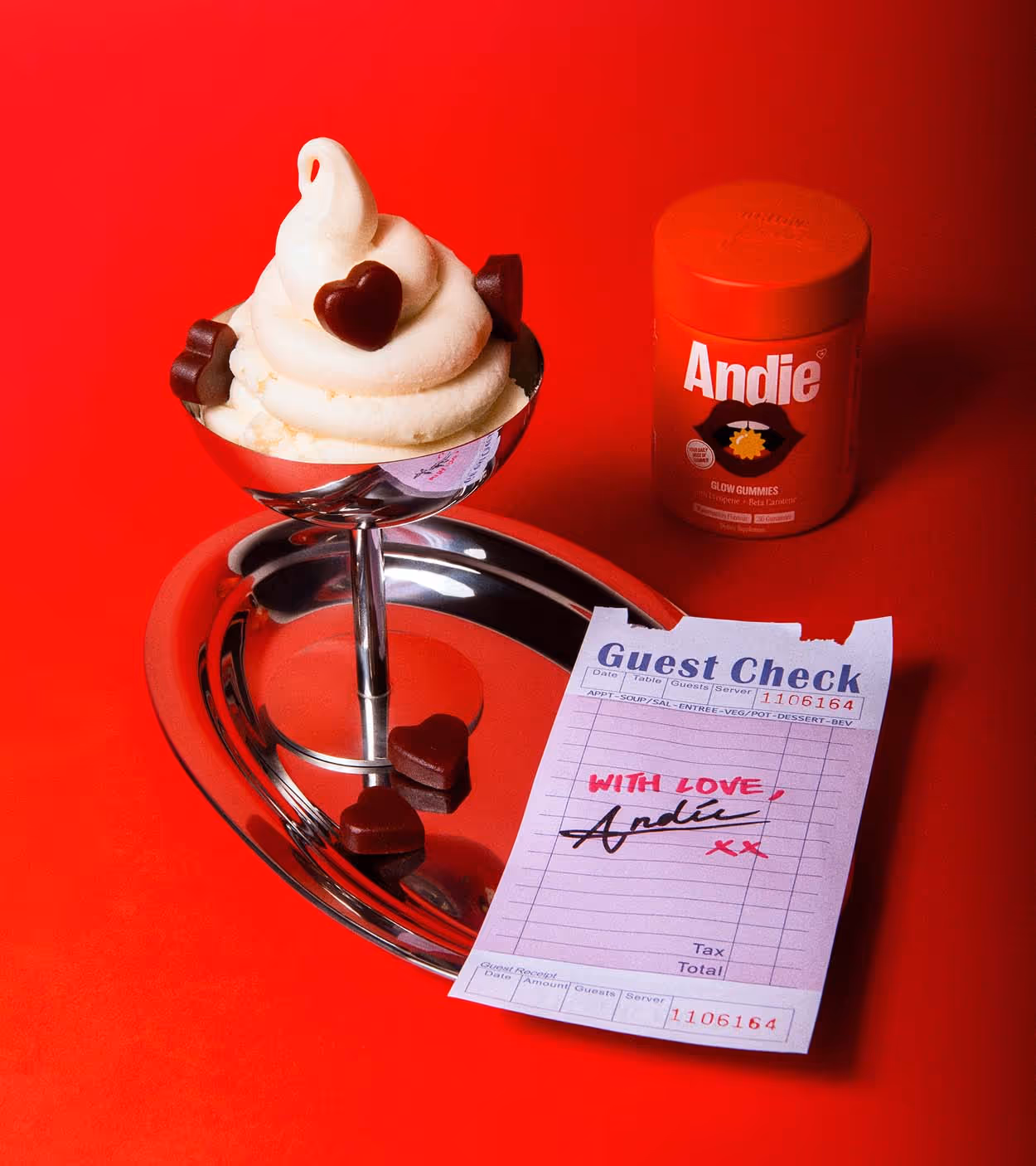 A silver dessert dish with soft serve topped with chocolate hearts, a red container of Andie Glow Gummies, and a guest check reading 'With love, Andie xx' on a reflective silver tray against a red background.