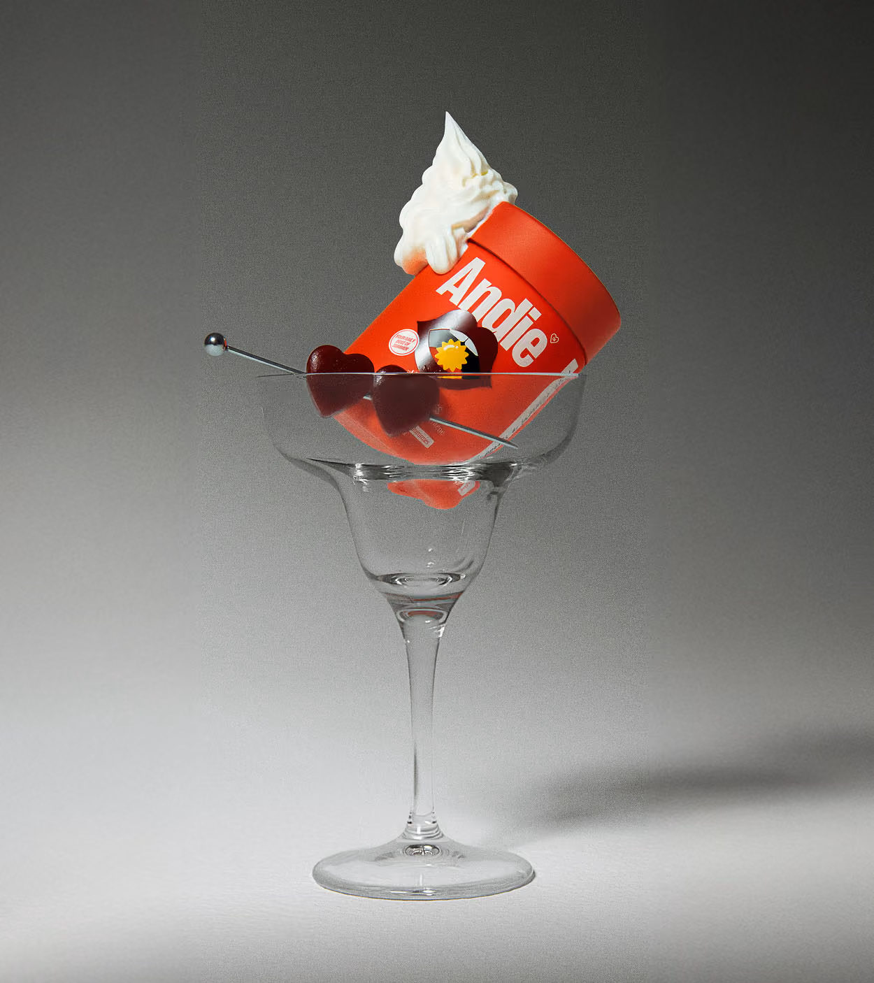 Red Andie product container with whipped topping inside a cocktail glass, garnished with two heart-shaped cherries on a skewer.